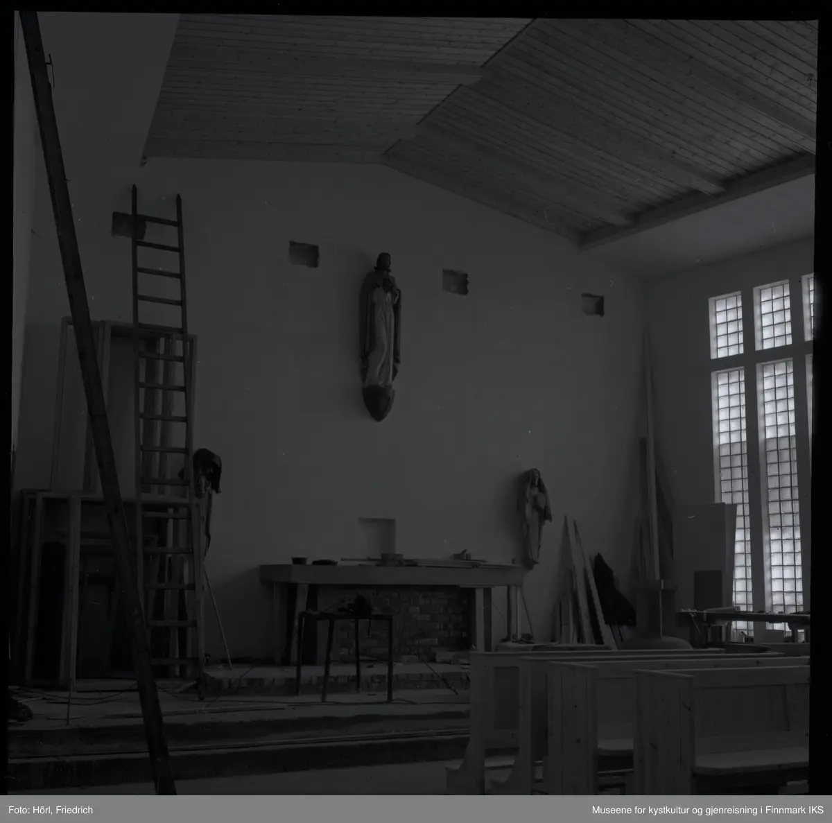 Interiøret til den katolske kirka St. Mikael i Hammerfest bygges i 1958. I motsetning til senere, er det montert en skulptur av Jesus Kristus ovenfor alteret. Senere ble det plassert et krusifiks der. Mosaikken på fronten av alteret som ble utført av Karl Manninger, er ikke kommet på plass enda. Til venstre for alteret er det montert en skulptur av Maria med barnet, til høyre vises Josef. Disse to skulpturene som er skjært av tre er tildekket med tepper. Foran alteret er det satt opp en bordsag. Til venstre i bilde står en lang stige og noen rammer lent til veggen, til høyre ved vinduene ser man en høvelbenk og mer byggemateriale. I forgrunnen til høyre står det kirkebenker.