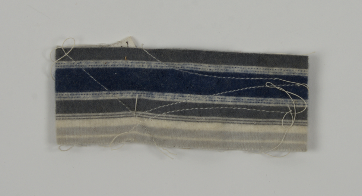 a) Symaskin av märket Husqvarna, modell V.S.4. Handdriven. Svartmålad på träplatta. Nersänkt låda med tillbehör. b) Huv av ekfaner. Handtag på toppen. c) Skyttel av stål. Rund, spetsig i ena änden, rak i den andra. Innehåller en spole med vit tråd. d) Fem spolar av stål. Rund stav med plattor i ändarna. Olika trådrester. e) Ledpressarfot (monterad på maskinen). f) Fållvikare 2 mm. g) Fållvikare 6 mm. h) Gansapparat. i) Rynkfot .j) Linjal. Längd 5,3 cm. k) Fem symaskinsnålar i olika grovlekar. Cellofanförpackning. l) Skruvnyckel. Typ lång platt nål, bakre delen försedd med runt hål. m) Skruvmejsel av stål. Fäste av svart bakelit. n) Borste av dubbelvriden ståltråd med en borste i ena änden. o) Skruvmejsel av stål. Fäste av svart bakelit. p) Provlapp av bomullsflanell. För kontroll av stygnställning. q) Provlapp av bomyllskypert. Till kontroll av stygnställning. r) Bruksanvisning. Tryckt broschyr på 30 sidor.