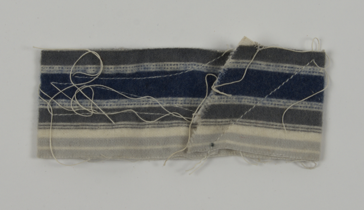 a) Symaskin av märket Husqvarna, modell V.S.4. Handdriven. Svartmålad på träplatta. Nersänkt låda med tillbehör. b) Huv av ekfaner. Handtag på toppen. c) Skyttel av stål. Rund, spetsig i ena änden, rak i den andra. Innehåller en spole med vit tråd. d) Fem spolar av stål. Rund stav med plattor i ändarna. Olika trådrester. e) Ledpressarfot (monterad på maskinen). f) Fållvikare 2 mm. g) Fållvikare 6 mm. h) Gansapparat. i) Rynkfot .j) Linjal. Längd 5,3 cm. k) Fem symaskinsnålar i olika grovlekar. Cellofanförpackning. l) Skruvnyckel. Typ lång platt nål, bakre delen försedd med runt hål. m) Skruvmejsel av stål. Fäste av svart bakelit. n) Borste av dubbelvriden ståltråd med en borste i ena änden. o) Skruvmejsel av stål. Fäste av svart bakelit. p) Provlapp av bomullsflanell. För kontroll av stygnställning. q) Provlapp av bomyllskypert. Till kontroll av stygnställning. r) Bruksanvisning. Tryckt broschyr på 30 sidor.