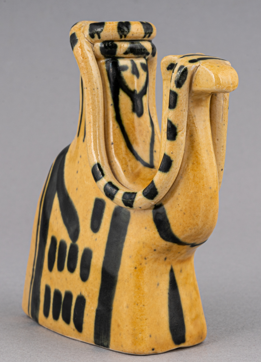 Figurin i lergods, kan även användas som ljusstake för julgransljus. Modell 6001, formgiven av Eva Jancke Björk under 1940- eller 1950-talet. Glasyr/dekor SV; curryfärgad bottenglasyr med svart dekor. Figurinen föreställer en arab sittandes på en kamel.