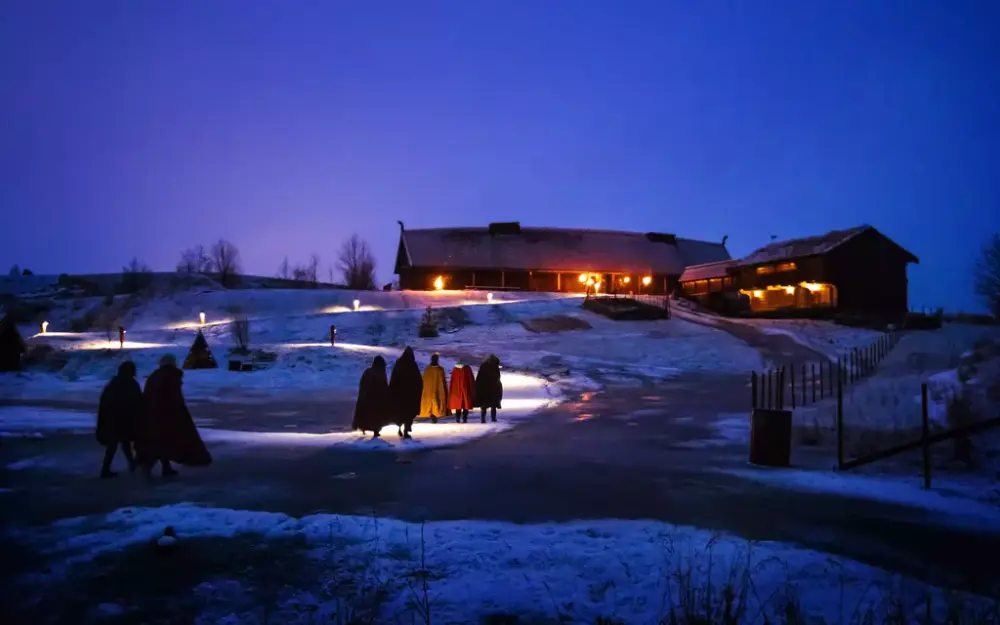 Mennesker kledt i vikingkapper går oppover en sti lyssatt med fakler opp mot en replika av et langhus fra vikingtiden