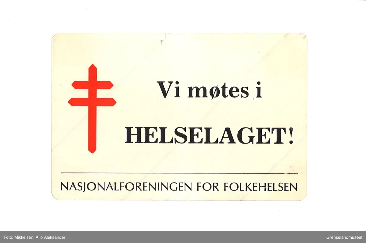 Hvit-beige klistrelappskilt hvor det står med svart skrift "Vi møtes i Helselaget!". Under den øverste teksten er det en linje og navnet til foreningen "Nasjonalforening for Folkehelsen." Helt til venstre på skiltet er symbolet til" Nasjonalforeningen for Folkehelsen", et rødt kors med to streker.
