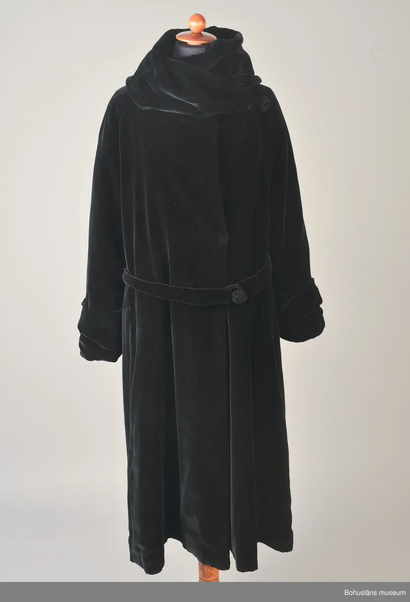 Kappa i svart sammet med vitt sidenfoder. Pellerinkrage och löst sittande skärp.