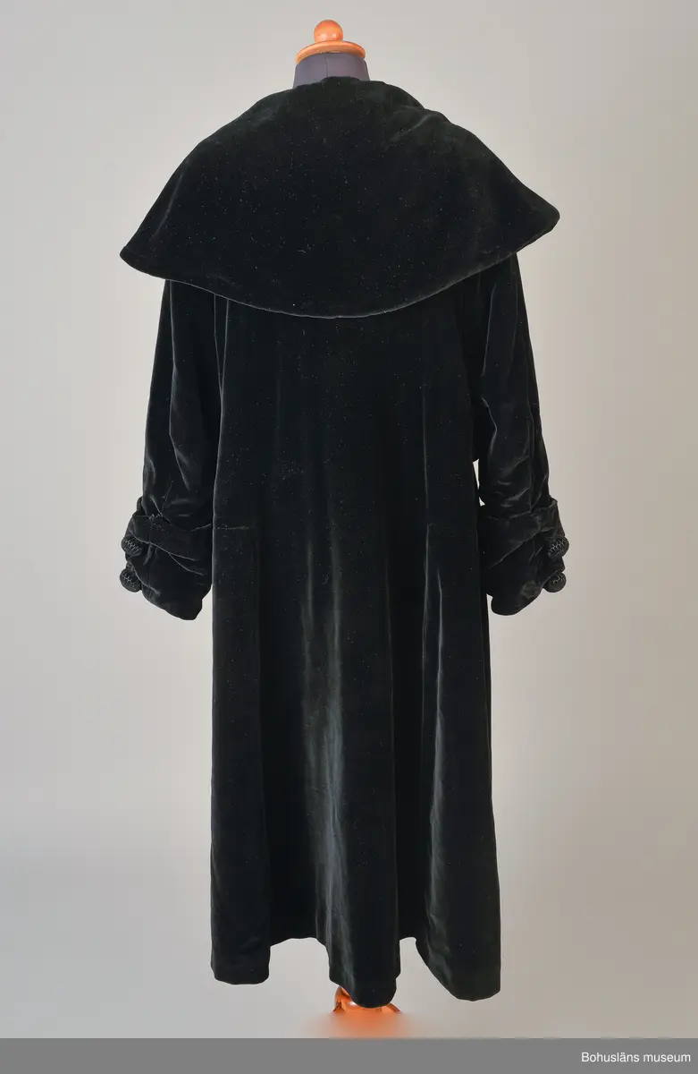 Kappa i svart sammet med vitt sidenfoder. Pellerinkrage och löst sittande skärp.