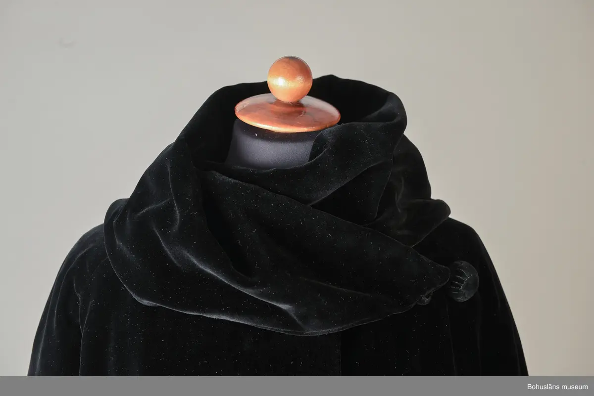 Kappa i svart sammet med vitt sidenfoder. Pellerinkrage och löst sittande skärp.