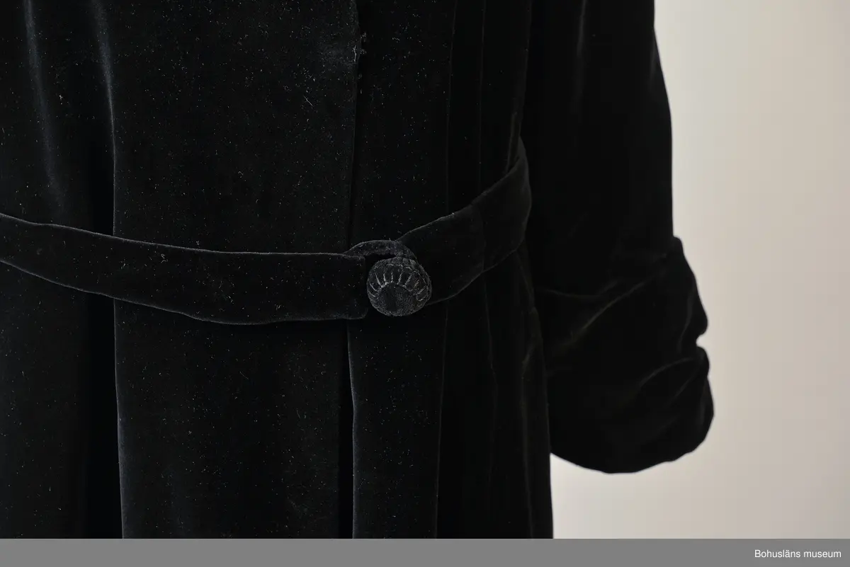 Kappa i svart sammet med vitt sidenfoder. Pellerinkrage och löst sittande skärp.