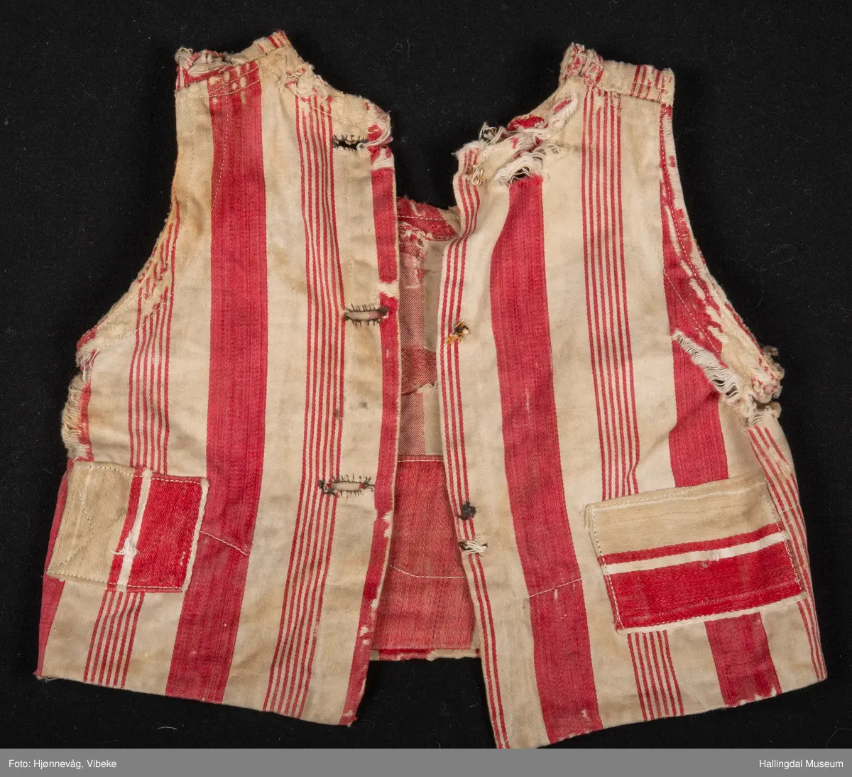 Trøye/vest uten ermer. Bomullstøy med brede røde og hvite striper. 2 pyntelommer, 3 knapphull. Knappene mangler. Stor bot i ryggen. Veldig slitt.