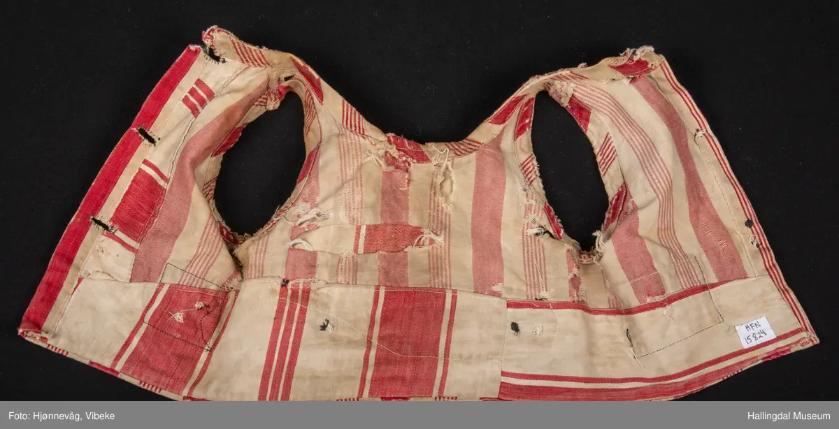 Trøye/vest uten ermer. Bomullstøy med brede røde og hvite striper. 2 pyntelommer, 3 knapphull. Knappene mangler. Stor bot i ryggen. Veldig slitt.