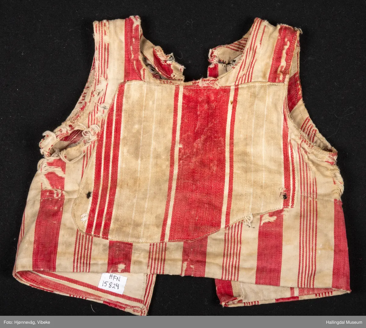 Trøye/vest uten ermer. Bomullstøy med brede røde og hvite striper. 2 pyntelommer, 3 knapphull. Knappene mangler. Stor bot i ryggen. Veldig slitt.