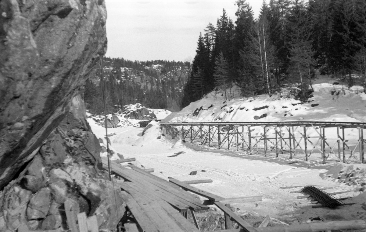 Fra bygginga av en ny dam ved Sølvstufossen, øverst i Ågårdselva i Tune i Østfold, som er en del av Nedre Glommas vestre løp. Anleggsarbeidet startet vinteren 1936, i en periode da det knapt rant vann i elveløpet. Fotografiet er tatt fra en gangbane, bygd som et trestillas langs det bratte og til dels overhengende berget langs søndre elvebredd. Til høyre ser vi en trallebane, som sto på trebukker langsmed nordre elvebredd. Den var antakelig bygd for å oppnå effektiv transport av redskaper og materialer til og fra byggeplassen. På isen, nederst i bildets høyre hjørne, lå det noe som kan se ut som jernstenger til armering av det murverket som skulle støpes. Ågårdselva er et cirka fem og en halv kilometer langt vassdrag som renner fra den nordvestre enden av Isnesfjorden (Vestvannet) i Nedre Glommas vestre løp, sørvestover gjennem berglendt terreng mot innsjøen Visterflo. Høydeforskjellen mellom Isnesfjorden og Visterflo er på bortimot 25 meter. Ågårdselva har tre fossefall, det øverste her ved Sølvstu, deretter ved Valbrekke og nederst ved Solli. Behovet for den dambygginga vi ser på dette fotografiet var forårsaket av selskapene Borregaards og Hafslunds inngrep i Glommas østre løp, hovedløpet, som i lavvannsperioder gjorde det svært vanskelig å få tømmer som skulle til bedrifter nedenfor Sarpsfossen via Mingevannet, Isnesfjorden og tømmertunnelen til Eidet gjennom det trange sundet ved Trøsken. Dette forsøkte man å løse ved å heve vannspeilet i Isnesfjorden med en dam som skulle plasseres 10-15 meter ovenfor den eksisterende dammen ved Sølvstufossen. Se mer informasjon under fanen «Opplysninger».