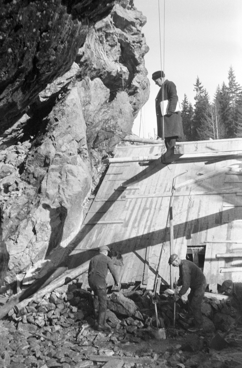 Fra bygginga av en ny dam ved Sølvstufossen, øverst i Ågårdselva i Tune i Østfold, som er en del av Nedre Glommas vestre løp. Anleggsarbeidet startet vinteren 1936, i en periode da det knapt rant vann i elveløpet. Fotografiet er tatt fra elveløpet, mot et skråplan av tre, sannsynligvis noe som etter hvert skulle bli et damløp. Nedenfor dette skråplanet var to arbeidere i aktivitet med spett og spade. På den øvre delen av planet sto lensebestyreren var Eidet, Hans Christian Skaiaa, kledd i svart vinterfrakk, med pelslue på hodet og dokumenter - antakerlig konstruksjonstegninger - under armen. Ågårdselva er et cirka fem og en halv kilometer langt vassdrag som renner fra den nordvestre enden av Isnesfjorden (Vestvannet) i Nedre Glommas vestre løp, sørvestover gjennem berglendt terreng mot innsjøen Visterflo. Høydeforskjellen mellom Isnesfjorden og Visterflo er på bortimot 25 meter. Ågårdselva har tre fossefall, det øverste her ved Sølvstu, deretter ved Valbrekke og nederst ved Solli. Behovet for den dambygginga vi ser på dette fotografiet var forårsaket av selskapene Borregaards og Hafslunds inngrep i Glommas østre løp, hovedløpet, som i lavvannsperioder gjorde det svært vanskelig å få tømmer som skulle til bedrifter nedenfor Sarpsfossen via Mingevannet, Isnesfjorden og tømmertunnelen til Eidet gjennom det trange sundet ved Trøsken. Dette forsøkte man å løse ved å heve vannspeilet i Isnesfjorden med en dam som skulle plasseres 10-15 meter ovenfor den eksisterende dammen ved Sølvstufossen. Se mer informasjon under fanen «Opplysninger».