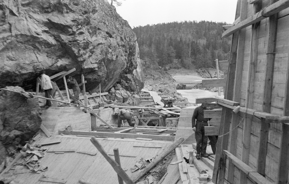 Fra bygginga av Sølvstudammen i Tune i Østfold i 1936. Denne dammen ligger i Ågårdselva, et cirka fem og en halv kilometer langt vassdrag som renner fra den nordvestre enden av Isnesfjorden (Vestvannet) i Nedre Glommas vestre løp, sørvestover gjennem berglendt terreng mot innsjøen Visterflo. Høydeforskjellen mellom Isnesfjorden og Visterflo er på bortimot 25 meter. Ågårdselva har tre fossefall, det øverste her ved Sølvstu, deretter ved Valbrekke og nederst ved Solli. Fotografiet er tatt gjennom det som skulle bli damløpet i Sølvstudammen, i medstrøms retning. Både fra elvebotnen mot damkrona og langs sidene var det bygd forskalinger av bord på et rammeverk av 2" X 2" og 4" X 4". Fem av forskalingssnekkerne er med på fotografiet. Noe høyere oppe ses et stillas med bordgang, hvor det sto ei trillebår. I bakgrunnen skimtes noe av elveleiet og en åskam med barskog.