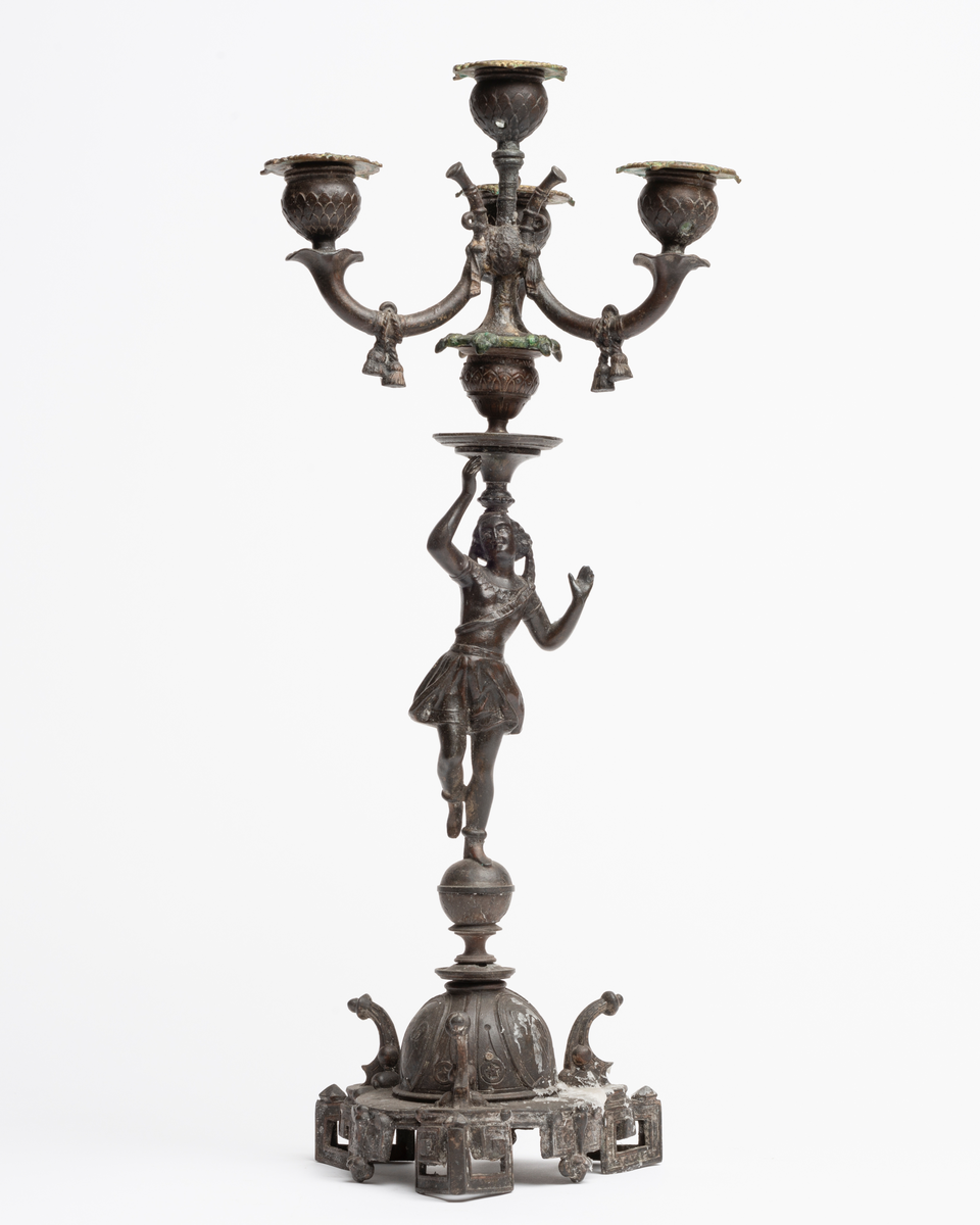 3-armet lysestake i metall. Skaftet består av en kvinnestate som balanserer på en kule. På hodet bærer hun selve lysholderen.