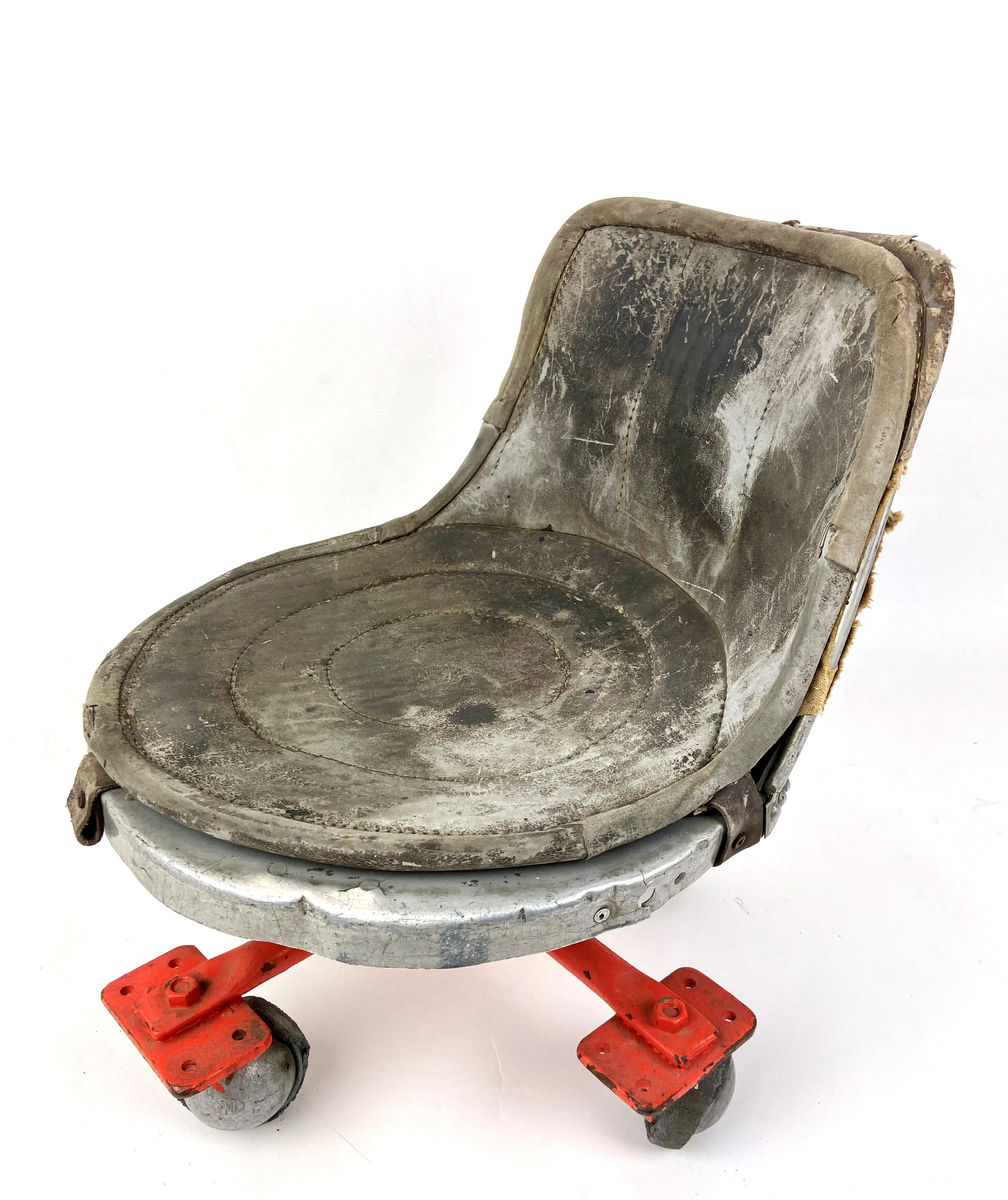 Flygplansstol, rund sits i skinn med kort ryggplatta. Stomme av metall. Flygplansstolen är omgjord till verkstadsstol med fyra hjul. Klädd med smärting på rayggplatta bak.