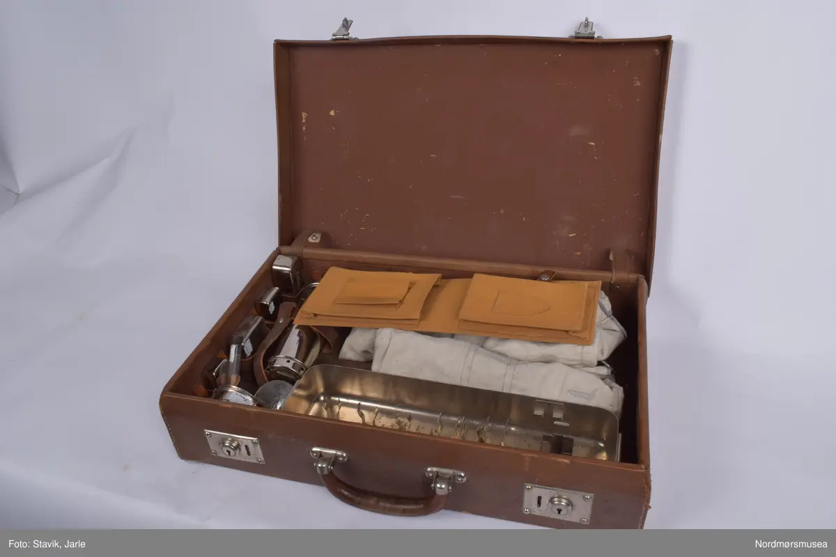 Koffert i brunt skinn.. Inneholder de fleste gjenstander man kan få bruk for ved fødsel. Full av utstyr a-å og aa-hh. Flere sylindre med diverse utstyr, noe løst utstyr, to tøyfuteraler for instrumenter.
a) Etui i rustfritt stål til børste merket Seifenbürste. Børsten i tre med krummet rygg. 10,5x3,7x3,4 cm. Børsten brukt til å skrubbe under neglene.
b) Etui til såpe i rustrfritt stål. Tomt, men lukter såpe. "Seife" på lokket. 8,2x4,2x2,5 cm.
c) Forniklet etui til oppbevaring av ampuller. Logo på lokket. Lokk hengslet, merket AESCULAP. 8x6,5x2,7 cm.
d) Sprøyte i forniklet etui med 2 spisser. "Original record" skrevet på lokket. 8x4x2 cm
e)Sylinderetui med lokk i begge ender. Hull i sidene på lokk og sylinder for lufting. 20,5 cm lang 7 cm diameter.
f) Sylinderlignende todelt sak. Ukjent funksjon. 8,3 cm diameter. 5,4 cm høyde.
g) 2 eterflasker 50g i brunt glass til bedøvelse oppbevart i kar i fødselskoffert.  "Eter til Narkose (Aether ad narcosin) N. sp. nr 2432. Tilsatt 0,001% DIEFENYLAMIN. Fabrikasjonsdatum 4. mar 1965." Prodsuert av A/S Den norske eterfabrikk. Målestreker med amrkering for 10.30 og 50 g.
h) Flat, rundt ekse i rustfritt stål.
i) Kar med utfoldbare føtter i rustrfitt stål til sterilisering med varmt vann.
j) Løslommer for obbevaring av instrumenter mm. Gult stivt tøy med solid skomakersøm.
m) Stort tøyfuteral i beige tøy med grå slitekanter. Trykknapper. Lommer og hemper inni med forskjellig fødselsutstyr. n-1 oppbevares i denne.
n) Simpsons fødselstang i rustfritt stål. I to deler for enkel sterilisering. Skeformede ender. Håndtaket har markerte fingergrept. Brukt til tangforløsning der fødselene får hjelp av en fødselstang i siste del av utdrivningsfasen. Dreier fosterhodet og trekker hodet gjennom fødselsåpningen. 35 cm lang.
p) Tang, arbortredskap i rustrfritt stål. Skarp saksaktig ende "til mutilering av foster i morsliv". Fjær på innsiden. Knapp for å holde tangen i stilling. Tangen er skap på utsiden i stedet for innsiden og spydformet når lukket. 27,5 lang, 4 cm langt blad.
q) Lang tang, sannsynligvis for "mutilering av foster i morsliv". Krummet ende, rillet på innsiden. Kan skrus sammen med skruer og vingemutter på enden. Kan deles i fire. 37 cm lang.
r) Pinsett, arteriepinsett til å klipe sammen blodårer med. 19,5 cm lang.
s) Nåleholder, sakslignede redskap for å holde suturnål. Haker for å låse den i stilling. 17 cm lang
t) Vaginalspekulum. Føres inn i skjeden for å holde den åpen under undersøkelser. Bøyd skaftende for bedre grep. Lik SU.3019u. 17cm lang,.
u)Vaginalspekulum ellers lik forrige bokstav.
v) Vaginalspekulum i rustfritt stål, føres inn i skjeden for å holde skjedeveggene åpne for undersøkelse. Fingergrept på skaftet med "avtrekker" til pekefinger for bedre grep. 22 cm lang.
w) Futeral i linstoff med tre trykknappet. Innvendig oppbevart forrskjellige instrumenter x-hh
x) Tang, kuletang, sakslignende instrument som brukes ved innsetting av spiral og gynekologiske undersøkelser. Rustfritt stål. 15 cm lang.
y) Lang kuletang, ellers lik x. 25 cm lang
z) Kuletang, lik y
æ) Korntang i rustfritt stål, rett skaft med mulighet for å låse den i sammenknepet stilling. 12,5 cm lang.
ø) Korntang lik æ
å) Sformet kateter (stikkes inn i urinøret for å tappe urin). Rustfritt stål.
aa) Spesialnål med mandreng. Mandreng er en nål som settes inn i hult rør eller kateter for at den lettere skal føres inn i organet som skal behandles. 9,5 cm lang.
bb) Spesialnål med mandreng 8 cm lang.
cc) Krum korntang i rustfritt stål, sakslignende med krummet ende. Kan låses i lukket stilling. 19 cm lang.
dd) Kirurgisaks med bøyd tipp i rustfritt stål. 14,5 cm lang.
ee) Korntang i rustfritt stål. Kan låses i lukket stilling.
ff) Tang, redskap for dilatasjon, utvide skjeden. Selvholdende slik at skjede forblir åpen. Håndtak rillet på hver ytterside av håndtaket. Mekanisme for å holde den åpen, justerbar. Krummet ende der tangendens bruksside er yttersiden. 16,5 cm lang.
hh) Rund eske i rustfritt stål til suturnåler. 3 nåler inni. Innskrift: AESCULAP. 6,5 cm diameter, 1 cm høy.
bb) 
