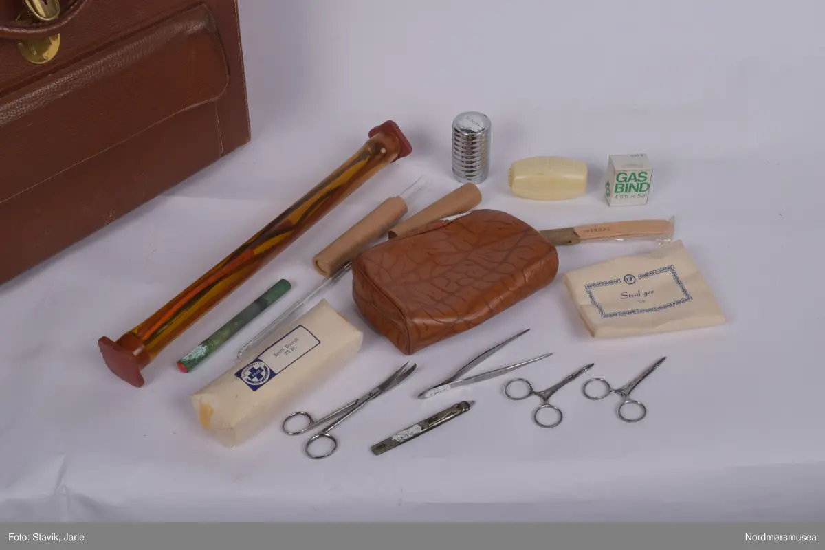 Legeveske i brukt skinn, åpning i toppen. Inneholder de fleste gjenstander man kan få bruk for ved sykebesøk. Dobbelt håndtak, lomme på utsiden kan lukkes med rein. Inni er hovedrom i bunnen med plastlommer med strikk øverst på en siden. Under denne to plastlommer med klaff. Et mindre skille av bunnen med skillevegg av gjenomsiktig plast. Inneholder numrene SU.3018a-3018s.
a) Etui for urinkatetere med fire katetre. Sylinder av gjennomsiktig plast med rød gummitopp i hver ende. 39 cm lang, 4 cm bred.
b) Sylindrisk metalletui i to deler for "smøreolje". Riller på siden fungerer som gjenger og dekorasjon. 78 cm langt, 4 cm diameter.
c) Todelt sylindrisk pappetui med reagensrør inni. 16,2 lang, røret 15,5 cm langt.
d) Termometer fra Villiers, merket ATOMIC. Liten glasshempe i toppen. 17 cm langt.
e) Tomt etui i messing med røde plastender. Irret.
f) Tomt etui i brunt skinn for blodtrykksapparat. Glidelås langs begges sider og topp. 17x12 cm.
g) Etui i eggehvit plast. Todelt med hengsling, inneholder staseslange produsert av Premeta Stauer. Brukes ved blodprøvetagning for å stramme rundt armen for å få fram blodårene.
m) Saks med krumme blader. 13 cm lang.
n) Skaplell med utfellbart blad. En skyvelås brukes for å hindre bladet i å folde seg sammen. Produsert av A. Rudds eftf. 14,5 cm lang utslått.
o) Anatomisk pinsett prdusert av Stille - Sverige. 13 cm langt.
p) To artieriepinsetter. Sakslignende pinsetter for å klipe igjen blodårer.
r) 4 pakker med steril suturtråd prod. av Davis & Beck Division American cyanid company. 1 pakke gasbind 4cmx5cm, 1 pk steril bomullsbandasje prod. av Beng & tegner & Co 25gr, 1 pk steril gasbind 1/4m, 1 stk trekantforkle 2,70.
s) 2 tungespatler i plast, 14,5 cm lang.