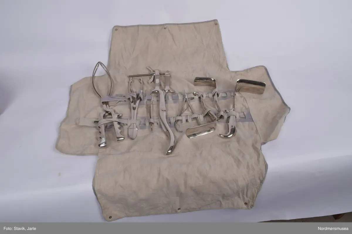 Koffert i brunt skinn.. Inneholder de fleste gjenstander man kan få bruk for ved fødsel. Full av utstyr a-å og aa-hh. Flere sylindre med diverse utstyr, noe løst utstyr, to tøyfuteraler for instrumenter.
a) Etui i rustfritt stål til børste merket Seifenbürste. Børsten i tre med krummet rygg. 10,5x3,7x3,4 cm. Børsten brukt til å skrubbe under neglene.
b) Etui til såpe i rustrfritt stål. Tomt, men lukter såpe. "Seife" på lokket. 8,2x4,2x2,5 cm.
c) Forniklet etui til oppbevaring av ampuller. Logo på lokket. Lokk hengslet, merket AESCULAP. 8x6,5x2,7 cm.
d) Sprøyte i forniklet etui med 2 spisser. "Original record" skrevet på lokket. 8x4x2 cm
e)Sylinderetui med lokk i begge ender. Hull i sidene på lokk og sylinder for lufting. 20,5 cm lang 7 cm diameter.
f) Sylinderlignende todelt sak. Ukjent funksjon. 8,3 cm diameter. 5,4 cm høyde.
g) 2 eterflasker 50g i brunt glass til bedøvelse oppbevart i kar i fødselskoffert.  "Eter til Narkose (Aether ad narcosin) N. sp. nr 2432. Tilsatt 0,001% DIEFENYLAMIN. Fabrikasjonsdatum 4. mar 1965." Prodsuert av A/S Den norske eterfabrikk. Målestreker med amrkering for 10.30 og 50 g.
h) Flat, rundt ekse i rustfritt stål.
i) Kar med utfoldbare føtter i rustrfitt stål til sterilisering med varmt vann.
j) Løslommer for obbevaring av instrumenter mm. Gult stivt tøy med solid skomakersøm.
m) Stort tøyfuteral i beige tøy med grå slitekanter. Trykknapper. Lommer og hemper inni med forskjellig fødselsutstyr. n-1 oppbevares i denne.
n) Simpsons fødselstang i rustfritt stål. I to deler for enkel sterilisering. Skeformede ender. Håndtaket har markerte fingergrept. Brukt til tangforløsning der fødselene får hjelp av en fødselstang i siste del av utdrivningsfasen. Dreier fosterhodet og trekker hodet gjennom fødselsåpningen. 35 cm lang.
p) Tang, arbortredskap i rustrfritt stål. Skarp saksaktig ende "til mutilering av foster i morsliv". Fjær på innsiden. Knapp for å holde tangen i stilling. Tangen er skap på utsiden i stedet for innsiden og spydformet når lukket. 27,5 lang, 4 cm langt blad.
q) Lang tang, sannsynligvis for "mutilering av foster i morsliv". Krummet ende, rillet på innsiden. Kan skrus sammen med skruer og vingemutter på enden. Kan deles i fire. 37 cm lang.
r) Pinsett, arteriepinsett til å klipe sammen blodårer med. 19,5 cm lang.
s) Nåleholder, sakslignede redskap for å holde suturnål. Haker for å låse den i stilling. 17 cm lang
t) Vaginalspekulum. Føres inn i skjeden for å holde den åpen under undersøkelser. Bøyd skaftende for bedre grep. Lik SU.3019u. 17cm lang,.
u)Vaginalspekulum ellers lik forrige bokstav.
v) Vaginalspekulum i rustfritt stål, føres inn i skjeden for å holde skjedeveggene åpne for undersøkelse. Fingergrept på skaftet med "avtrekker" til pekefinger for bedre grep. 22 cm lang.
w) Futeral i linstoff med tre trykknappet. Innvendig oppbevart forrskjellige instrumenter x-hh
x) Tang, kuletang, sakslignende instrument som brukes ved innsetting av spiral og gynekologiske undersøkelser. Rustfritt stål. 15 cm lang.
y) Lang kuletang, ellers lik x. 25 cm lang
z) Kuletang, lik y
æ) Korntang i rustfritt stål, rett skaft med mulighet for å låse den i sammenknepet stilling. 12,5 cm lang.
ø) Korntang lik æ
å) Sformet kateter (stikkes inn i urinøret for å tappe urin). Rustfritt stål.
aa) Spesialnål med mandreng. Mandreng er en nål som settes inn i hult rør eller kateter for at den lettere skal føres inn i organet som skal behandles. 9,5 cm lang.
bb) Spesialnål med mandreng 8 cm lang.
cc) Krum korntang i rustfritt stål, sakslignende med krummet ende. Kan låses i lukket stilling. 19 cm lang.
dd) Kirurgisaks med bøyd tipp i rustfritt stål. 14,5 cm lang.
ee) Korntang i rustfritt stål. Kan låses i lukket stilling.
ff) Tang, redskap for dilatasjon, utvide skjeden. Selvholdende slik at skjede forblir åpen. Håndtak rillet på hver ytterside av håndtaket. Mekanisme for å holde den åpen, justerbar. Krummet ende der tangendens bruksside er yttersiden. 16,5 cm lang.
hh) Rund eske i rustfritt stål til suturnåler. 3 nåler inni. Innskrift: AESCULAP. 6,5 cm diameter, 1 cm høy.
bb) 
