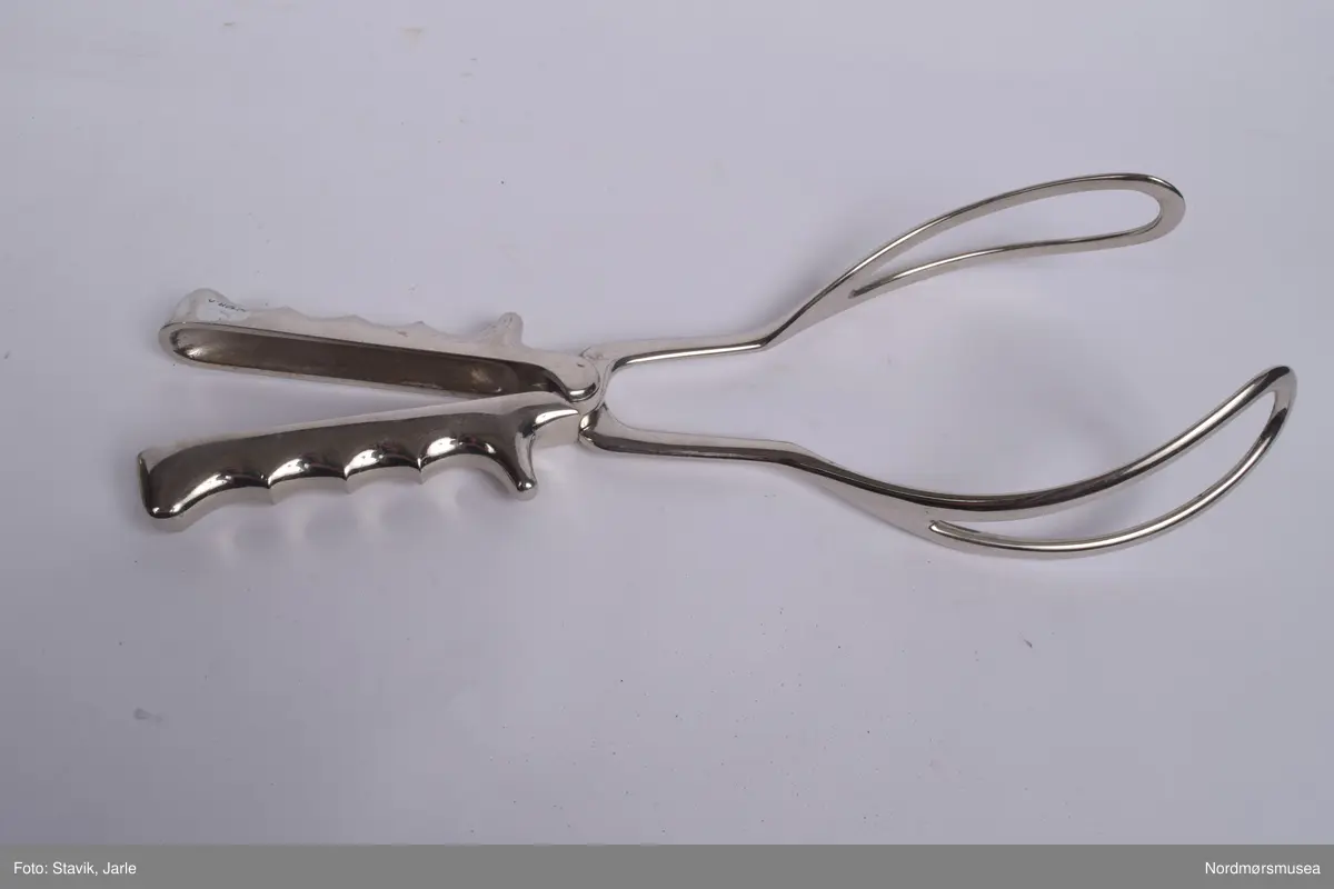 Koffert i brunt skinn.. Inneholder de fleste gjenstander man kan få bruk for ved fødsel. Full av utstyr a-å og aa-hh. Flere sylindre med diverse utstyr, noe løst utstyr, to tøyfuteraler for instrumenter.
a) Etui i rustfritt stål til børste merket Seifenbürste. Børsten i tre med krummet rygg. 10,5x3,7x3,4 cm. Børsten brukt til å skrubbe under neglene.
b) Etui til såpe i rustrfritt stål. Tomt, men lukter såpe. "Seife" på lokket. 8,2x4,2x2,5 cm.
c) Forniklet etui til oppbevaring av ampuller. Logo på lokket. Lokk hengslet, merket AESCULAP. 8x6,5x2,7 cm.
d) Sprøyte i forniklet etui med 2 spisser. "Original record" skrevet på lokket. 8x4x2 cm
e)Sylinderetui med lokk i begge ender. Hull i sidene på lokk og sylinder for lufting. 20,5 cm lang 7 cm diameter.
f) Sylinderlignende todelt sak. Ukjent funksjon. 8,3 cm diameter. 5,4 cm høyde.
g) 2 eterflasker 50g i brunt glass til bedøvelse oppbevart i kar i fødselskoffert.  "Eter til Narkose (Aether ad narcosin) N. sp. nr 2432. Tilsatt 0,001% DIEFENYLAMIN. Fabrikasjonsdatum 4. mar 1965." Prodsuert av A/S Den norske eterfabrikk. Målestreker med amrkering for 10.30 og 50 g.
h) Flat, rundt ekse i rustfritt stål.
i) Kar med utfoldbare føtter i rustrfitt stål til sterilisering med varmt vann.
j) Løslommer for obbevaring av instrumenter mm. Gult stivt tøy med solid skomakersøm.
m) Stort tøyfuteral i beige tøy med grå slitekanter. Trykknapper. Lommer og hemper inni med forskjellig fødselsutstyr. n-1 oppbevares i denne.
n) Simpsons fødselstang i rustfritt stål. I to deler for enkel sterilisering. Skeformede ender. Håndtaket har markerte fingergrept. Brukt til tangforløsning der fødselene får hjelp av en fødselstang i siste del av utdrivningsfasen. Dreier fosterhodet og trekker hodet gjennom fødselsåpningen. 35 cm lang.
p) Tang, arbortredskap i rustrfritt stål. Skarp saksaktig ende "til mutilering av foster i morsliv". Fjær på innsiden. Knapp for å holde tangen i stilling. Tangen er skap på utsiden i stedet for innsiden og spydformet når lukket. 27,5 lang, 4 cm langt blad.
q) Lang tang, sannsynligvis for "mutilering av foster i morsliv". Krummet ende, rillet på innsiden. Kan skrus sammen med skruer og vingemutter på enden. Kan deles i fire. 37 cm lang.
r) Pinsett, arteriepinsett til å klipe sammen blodårer med. 19,5 cm lang.
s) Nåleholder, sakslignede redskap for å holde suturnål. Haker for å låse den i stilling. 17 cm lang
t) Vaginalspekulum. Føres inn i skjeden for å holde den åpen under undersøkelser. Bøyd skaftende for bedre grep. Lik SU.3019u. 17cm lang,.
u)Vaginalspekulum ellers lik forrige bokstav.
v) Vaginalspekulum i rustfritt stål, føres inn i skjeden for å holde skjedeveggene åpne for undersøkelse. Fingergrept på skaftet med "avtrekker" til pekefinger for bedre grep. 22 cm lang.
w) Futeral i linstoff med tre trykknappet. Innvendig oppbevart forrskjellige instrumenter x-hh
x) Tang, kuletang, sakslignende instrument som brukes ved innsetting av spiral og gynekologiske undersøkelser. Rustfritt stål. 15 cm lang.
y) Lang kuletang, ellers lik x. 25 cm lang
z) Kuletang, lik y
æ) Korntang i rustfritt stål, rett skaft med mulighet for å låse den i sammenknepet stilling. 12,5 cm lang.
ø) Korntang lik æ
å) Sformet kateter (stikkes inn i urinøret for å tappe urin). Rustfritt stål.
aa) Spesialnål med mandreng. Mandreng er en nål som settes inn i hult rør eller kateter for at den lettere skal føres inn i organet som skal behandles. 9,5 cm lang.
bb) Spesialnål med mandreng 8 cm lang.
cc) Krum korntang i rustfritt stål, sakslignende med krummet ende. Kan låses i lukket stilling. 19 cm lang.
dd) Kirurgisaks med bøyd tipp i rustfritt stål. 14,5 cm lang.
ee) Korntang i rustfritt stål. Kan låses i lukket stilling.
ff) Tang, redskap for dilatasjon, utvide skjeden. Selvholdende slik at skjede forblir åpen. Håndtak rillet på hver ytterside av håndtaket. Mekanisme for å holde den åpen, justerbar. Krummet ende der tangendens bruksside er yttersiden. 16,5 cm lang.
hh) Rund eske i rustfritt stål til suturnåler. 3 nåler inni. Innskrift: AESCULAP. 6,5 cm diameter, 1 cm høy.
bb) 
