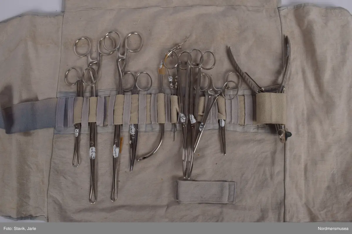 Koffert i brunt skinn.. Inneholder de fleste gjenstander man kan få bruk for ved fødsel. Full av utstyr a-å og aa-hh. Flere sylindre med diverse utstyr, noe løst utstyr, to tøyfuteraler for instrumenter.
a) Etui i rustfritt stål til børste merket Seifenbürste. Børsten i tre med krummet rygg. 10,5x3,7x3,4 cm. Børsten brukt til å skrubbe under neglene.
b) Etui til såpe i rustrfritt stål. Tomt, men lukter såpe. "Seife" på lokket. 8,2x4,2x2,5 cm.
c) Forniklet etui til oppbevaring av ampuller. Logo på lokket. Lokk hengslet, merket AESCULAP. 8x6,5x2,7 cm.
d) Sprøyte i forniklet etui med 2 spisser. "Original record" skrevet på lokket. 8x4x2 cm
e)Sylinderetui med lokk i begge ender. Hull i sidene på lokk og sylinder for lufting. 20,5 cm lang 7 cm diameter.
f) Sylinderlignende todelt sak. Ukjent funksjon. 8,3 cm diameter. 5,4 cm høyde.
g) 2 eterflasker 50g i brunt glass til bedøvelse oppbevart i kar i fødselskoffert.  "Eter til Narkose (Aether ad narcosin) N. sp. nr 2432. Tilsatt 0,001% DIEFENYLAMIN. Fabrikasjonsdatum 4. mar 1965." Prodsuert av A/S Den norske eterfabrikk. Målestreker med amrkering for 10.30 og 50 g.
h) Flat, rundt ekse i rustfritt stål.
i) Kar med utfoldbare føtter i rustrfitt stål til sterilisering med varmt vann.
j) Løslommer for obbevaring av instrumenter mm. Gult stivt tøy med solid skomakersøm.
m) Stort tøyfuteral i beige tøy med grå slitekanter. Trykknapper. Lommer og hemper inni med forskjellig fødselsutstyr. n-1 oppbevares i denne.
n) Simpsons fødselstang i rustfritt stål. I to deler for enkel sterilisering. Skeformede ender. Håndtaket har markerte fingergrept. Brukt til tangforløsning der fødselene får hjelp av en fødselstang i siste del av utdrivningsfasen. Dreier fosterhodet og trekker hodet gjennom fødselsåpningen. 35 cm lang.
p) Tang, arbortredskap i rustrfritt stål. Skarp saksaktig ende "til mutilering av foster i morsliv". Fjær på innsiden. Knapp for å holde tangen i stilling. Tangen er skap på utsiden i stedet for innsiden og spydformet når lukket. 27,5 lang, 4 cm langt blad.
q) Lang tang, sannsynligvis for "mutilering av foster i morsliv". Krummet ende, rillet på innsiden. Kan skrus sammen med skruer og vingemutter på enden. Kan deles i fire. 37 cm lang.
r) Pinsett, arteriepinsett til å klipe sammen blodårer med. 19,5 cm lang.
s) Nåleholder, sakslignede redskap for å holde suturnål. Haker for å låse den i stilling. 17 cm lang
t) Vaginalspekulum. Føres inn i skjeden for å holde den åpen under undersøkelser. Bøyd skaftende for bedre grep. Lik SU.3019u. 17cm lang,.
u)Vaginalspekulum ellers lik forrige bokstav.
v) Vaginalspekulum i rustfritt stål, føres inn i skjeden for å holde skjedeveggene åpne for undersøkelse. Fingergrept på skaftet med "avtrekker" til pekefinger for bedre grep. 22 cm lang.
w) Futeral i linstoff med tre trykknappet. Innvendig oppbevart forrskjellige instrumenter x-hh
x) Tang, kuletang, sakslignende instrument som brukes ved innsetting av spiral og gynekologiske undersøkelser. Rustfritt stål. 15 cm lang.
y) Lang kuletang, ellers lik x. 25 cm lang
z) Kuletang, lik y
æ) Korntang i rustfritt stål, rett skaft med mulighet for å låse den i sammenknepet stilling. 12,5 cm lang.
ø) Korntang lik æ
å) Sformet kateter (stikkes inn i urinøret for å tappe urin). Rustfritt stål.
aa) Spesialnål med mandreng. Mandreng er en nål som settes inn i hult rør eller kateter for at den lettere skal føres inn i organet som skal behandles. 9,5 cm lang.
bb) Spesialnål med mandreng 8 cm lang.
cc) Krum korntang i rustfritt stål, sakslignende med krummet ende. Kan låses i lukket stilling. 19 cm lang.
dd) Kirurgisaks med bøyd tipp i rustfritt stål. 14,5 cm lang.
ee) Korntang i rustfritt stål. Kan låses i lukket stilling.
ff) Tang, redskap for dilatasjon, utvide skjeden. Selvholdende slik at skjede forblir åpen. Håndtak rillet på hver ytterside av håndtaket. Mekanisme for å holde den åpen, justerbar. Krummet ende der tangendens bruksside er yttersiden. 16,5 cm lang.
hh) Rund eske i rustfritt stål til suturnåler. 3 nåler inni. Innskrift: AESCULAP. 6,5 cm diameter, 1 cm høy.
bb) 
