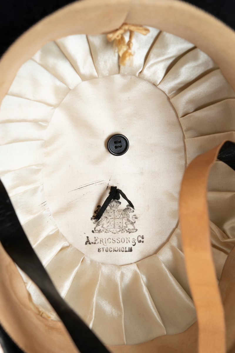 Paradhatt av bikorn-typ, även kallad båthatt, modell 1854 (m/1854). Svart felb på pressad papp. Gulddragarbete på hattens hörn och sida (agraff - bandkokard, dubbelsnöre och knapp/krönt löv). Strutsplym längs brättenas överkanter. Foder av vitt siden, svettrem och hakband av lackskinn.