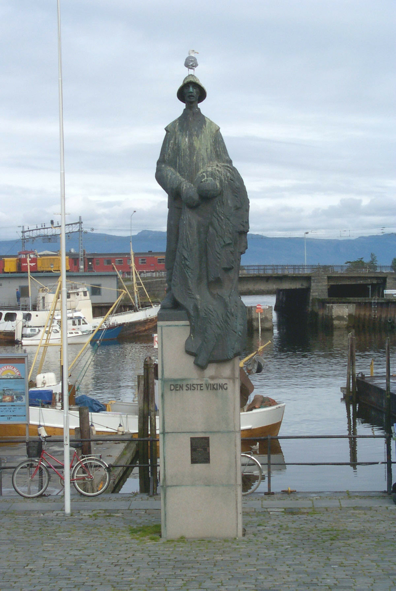 Helfigur på sokkel. En fisker som står med sitt fiskegarn i favnen. Antrukket i lang arbeidsfrakk og sydvest på hodet.