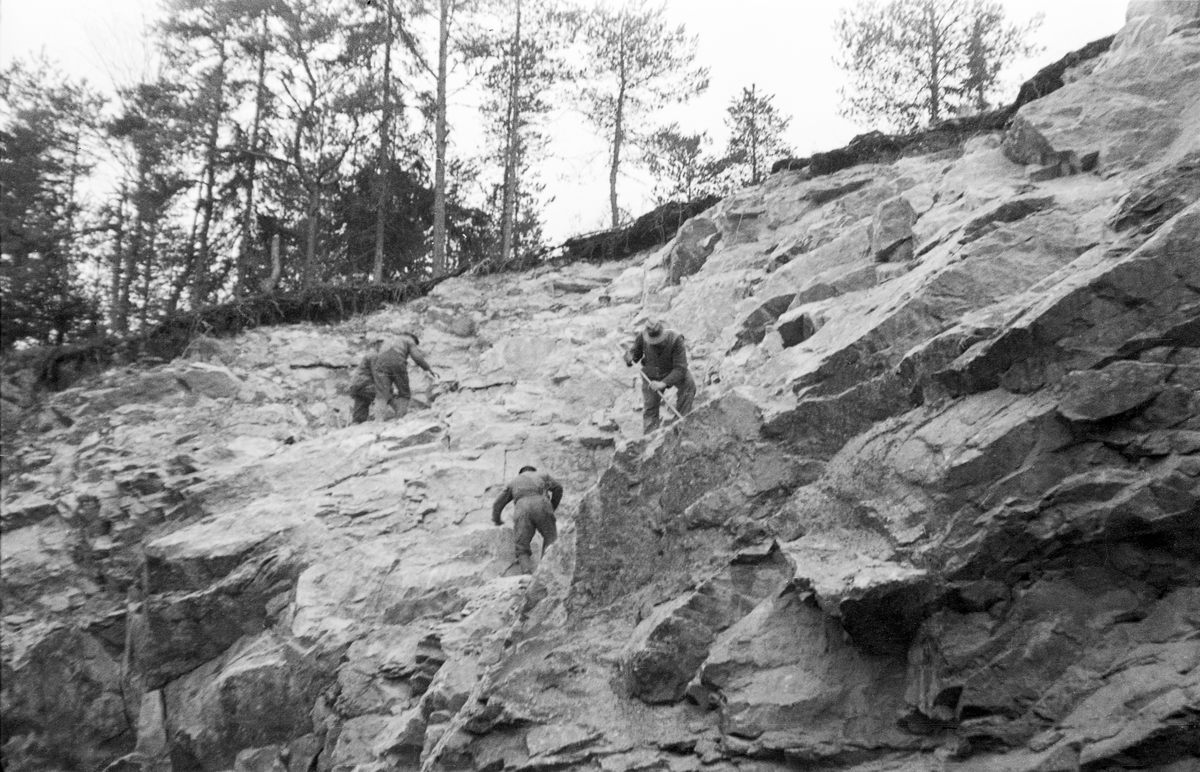 Fra bygginga av Sølvstudammen i Tune i Østfold i 1936. Fotografiet er tatt mot en bergskråning der det ble sprengt løs stein med tanke på oppfylling av underlaget for styrtgolvet under dammen. Tre menn arbeider i berget. En av dem står med spett og forsøker åpenbart å løsne stein.  Øverst på bakkekammen skimtes et tynt jordskikt med furuskog. Sølvstudammen ligger i Ågårdselva, et cirka fem og en halv kilometer langt vassdrag som renner fra den nordvestre enden av Isnesfjorden (Vestvannet) i Nedre Glommas vestre løp, sørvestover gjennem berglendt terreng mot innsjøen Visterflo. Høydeforskjellen mellom Isnesfjorden og Visterflo er på bortimot 25 meter. Ågårdselva har tre fossefall, det øverste her ved Sølvstu, deretter ved Valbrekke og nederst ved Solli. Behovet for den dambygginga vi ser på dette fotografiet var forårsaket av selskapene Borregaards og Hafslunds inngrep i Glommas østre løp, hovedløpet, som gjorde det svært vanskelig å få tømmer som skulle til bedrifter nedenfor Sarpsfossen via Mingevannet, Isnesfjorden og tømmertunnelen til Eidet gjennom det trange sundet ved Trøsken (mellom Mingevannet og Isnesfjorden) i lavvannsperioder. Dette forsøkte man å løse ved å heve vannspeilet i Isnesfjorden med en dam som skulle plasseres 10-15 meter ovenfor den eksisterende dammen ved Sølvstufossen. Se mer informasjon under fanen «Opplysninger».