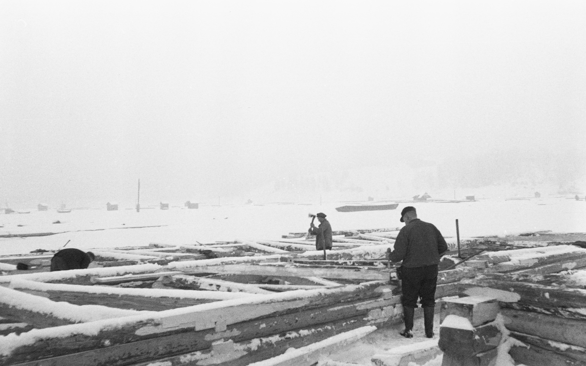 Bygging av fagverkslenser eller stivlenser til lenseanlegget på Glennetangen i Nedre Glomma, som her var grense mellom Skiptvet og Varteig kommuner, vinteren 1937. Dette var flytende stengsler som skulle holde seg rette sjøl om de ble utsatt for stort trykk, og som i mange tilfeller også skulle bære gangbaner som kunne tjene som arbeidsplattformer for fløterne. I dette tilfellet var lenseelementene bygd som rammer med tre-fire omfar med tømmer, avstivet med innvendige skråband i cirka 45 graders vinkel. Her ble komponentene bygd på isen vinterstid, men så snart isen smeltet ble de til flytende komponenter som kunne kjedes sammen i det som var i ferd med å bli et nytt sorterings- og moseanlegg (bunting av tømmer). Dette fotografiet viser tømmermennene i arbeid.