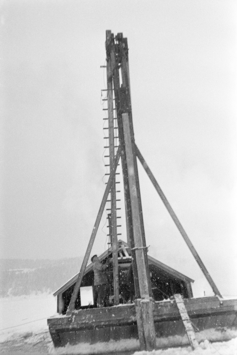 Pæletårn eller pælebukk på flåte. Fotografiet er tatt ved Glennetangen i Nedre Glomma, som her var grense mellom Skiptvet og Varteig kommuner. Fotografiet ble tatt vinteren 1937, da det ble bygd et nytt lenseanlegg her, hvor det tømmeret som skulle gå i Glommas østre løp, mot Sarpsborg, ble skilt fra det som via det vestre løpet skulle til bedrifter i Fredrikstad-distriktet og til trefordlingsanlegg andre steder ved Oslofjorden. De flytende stengslene i lenseanlegget måtte ha faste anleggspunkter i form av pæler, også kalt dopper eller duc d’alber. Disse ble drevet loddrett fire til seks meter ned i elvebotnen med rotendene nedovervendt. Pælestokkene måtte være lange nok til at toppendene stakk godt opp over vannspeilet. Her skulle diameteren være minst 15 centimeter, gjerne større. Pælene ble drevet ned i elvebotnen ved hjelp av et pæletårn – et høyt stativ med ei trinse i toppen. Over denne trinsa gikk det et tau eller en vaier, og i enden av denne var det et tungt lodd – i dette tilfellet en grov, rettvokst tømmerstokk. Ved å heve denne stokken, for deretter å la den falle ned på toppenden av pålen, gang etter gang, fikk man den etter hvert til å stå godt nok. Pælinga ble vanligvis utført vinterstid, ofte med pælebukken stående direkte på tjukk is. Her var isen antakelig for tynn og usikker, så det ble brukt en pælebukk som var montert i akterenden på en bred og stabil pram.