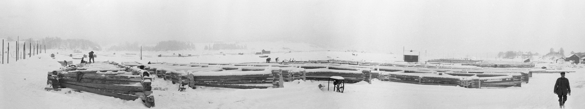 Bygging av fagverkslenser eller stivlenser til lenseanlegget på Glennetangen i Nedre Glomma, som her var grense mellom Skiptvet og Varteig kommuner, vinteren 1937. Dette var flytende stengsler som skulle holde seg rette sjøl om de ble utsatt for stort trykk, og som i mange tilfeller også skulle bære gangbaner som kunne tjene som arbeidsplattformer for fløterne. I dette tilfellet var lenseelementene bygd som rammer med tre-fire omfar med tømmer, avstivet med innvendige skråband i cirka 45 graders vinkel. Her ble komponentene bygd på isen vinterstid, men så snart isen smeltet ble de til flytende komponenter som kunne kjedes sammen i det som var i ferd med å bli et nytt sorterings- og moseanlegg (bunting av tømmer). Dette fotografiet viser tømmermennene i arbeid.