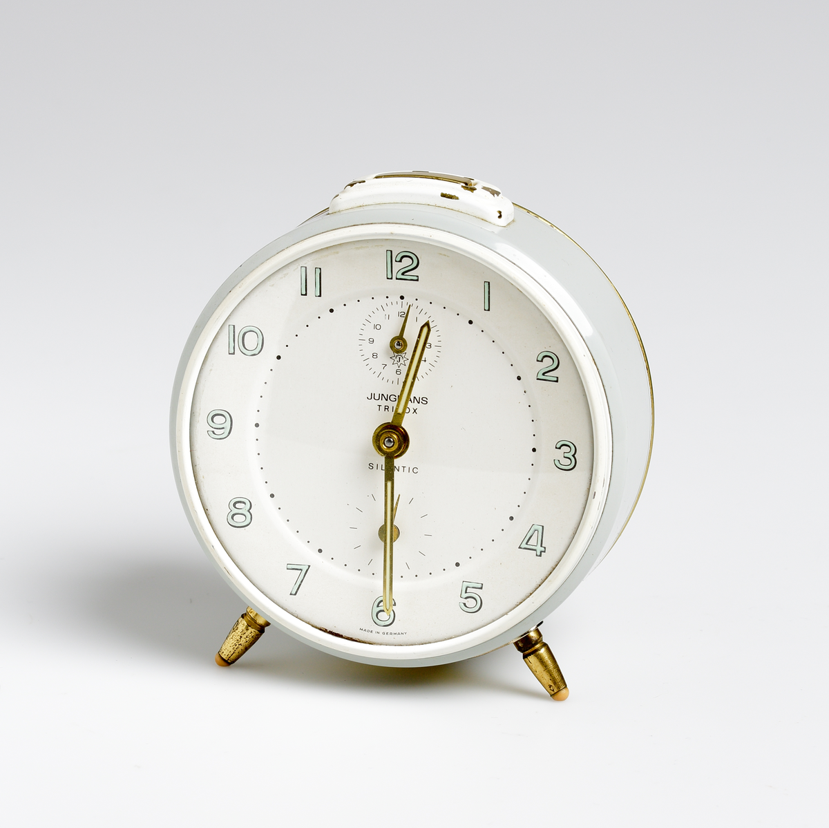Väckarklocka i grå- och vitlackad metall. Under urtavlan två mässingfötter med gummitassar. Vit urtavla med gröna självlysande siffror. Mässingvisare med självlysande grön färg. Text på urtavlan: "JUNGHANS TRIVOX // SILENTIC // MADE IN GERMANY". Vit etikett på baksidan: "A.B. KVARNSTRÖM OSKARSHAMN // IMPORT".