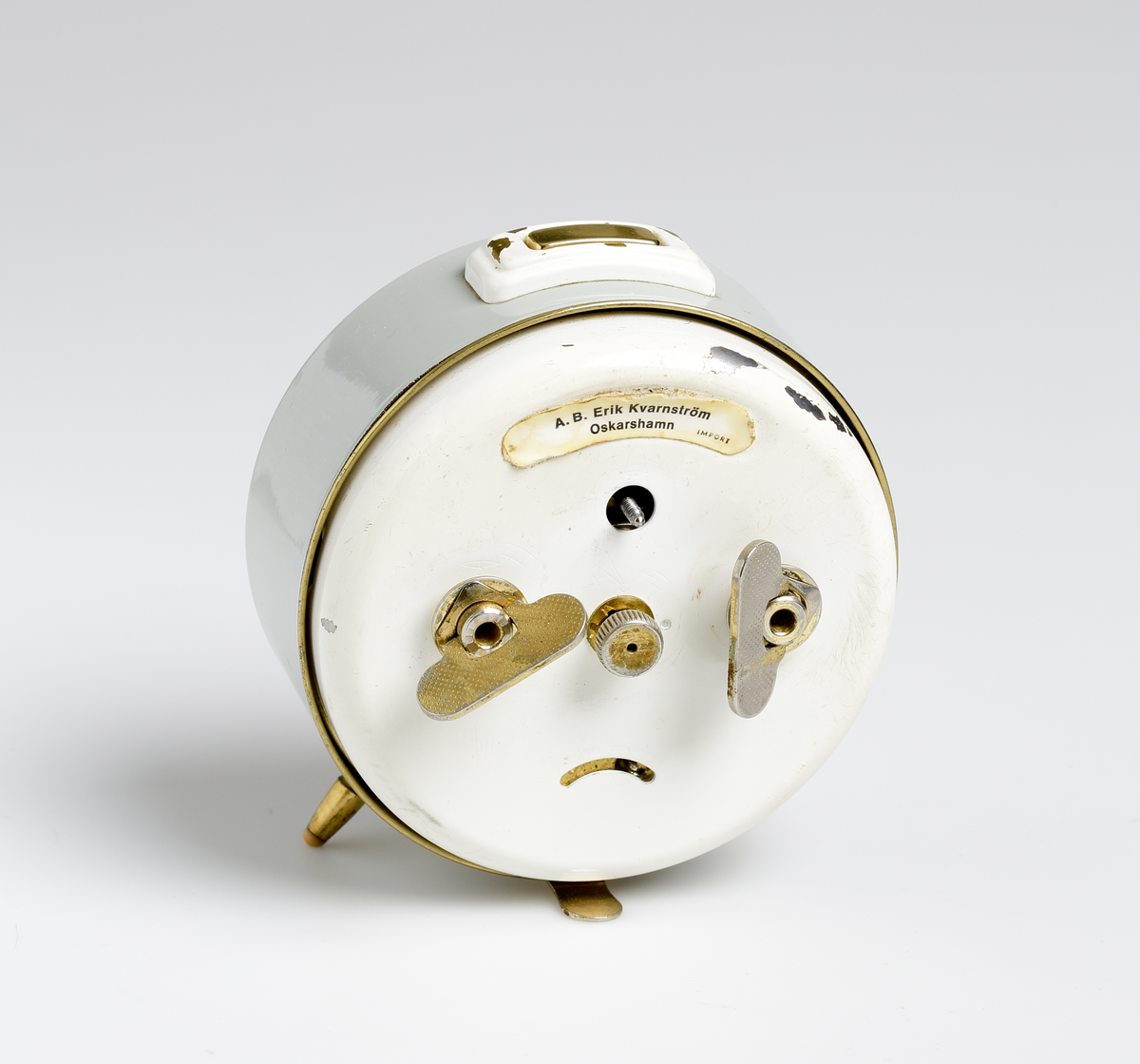 Väckarklocka i grå- och vitlackad metall. Under urtavlan två mässingfötter med gummitassar. Vit urtavla med gröna självlysande siffror. Mässingvisare med självlysande grön färg. Text på urtavlan: "JUNGHANS TRIVOX // SILENTIC // MADE IN GERMANY". Vit etikett på baksidan: "A.B. KVARNSTRÖM OSKARSHAMN // IMPORT".