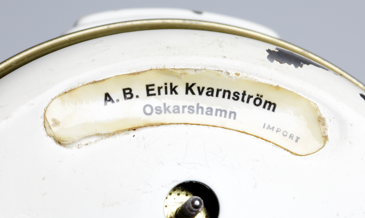 Väckarklocka i grå- och vitlackad metall. Under urtavlan två mässingfötter med gummitassar. Vit urtavla med gröna självlysande siffror. Mässingvisare med självlysande grön färg. Text på urtavlan: "JUNGHANS TRIVOX // SILENTIC // MADE IN GERMANY". Vit etikett på baksidan: "A.B. KVARNSTRÖM OSKARSHAMN // IMPORT".