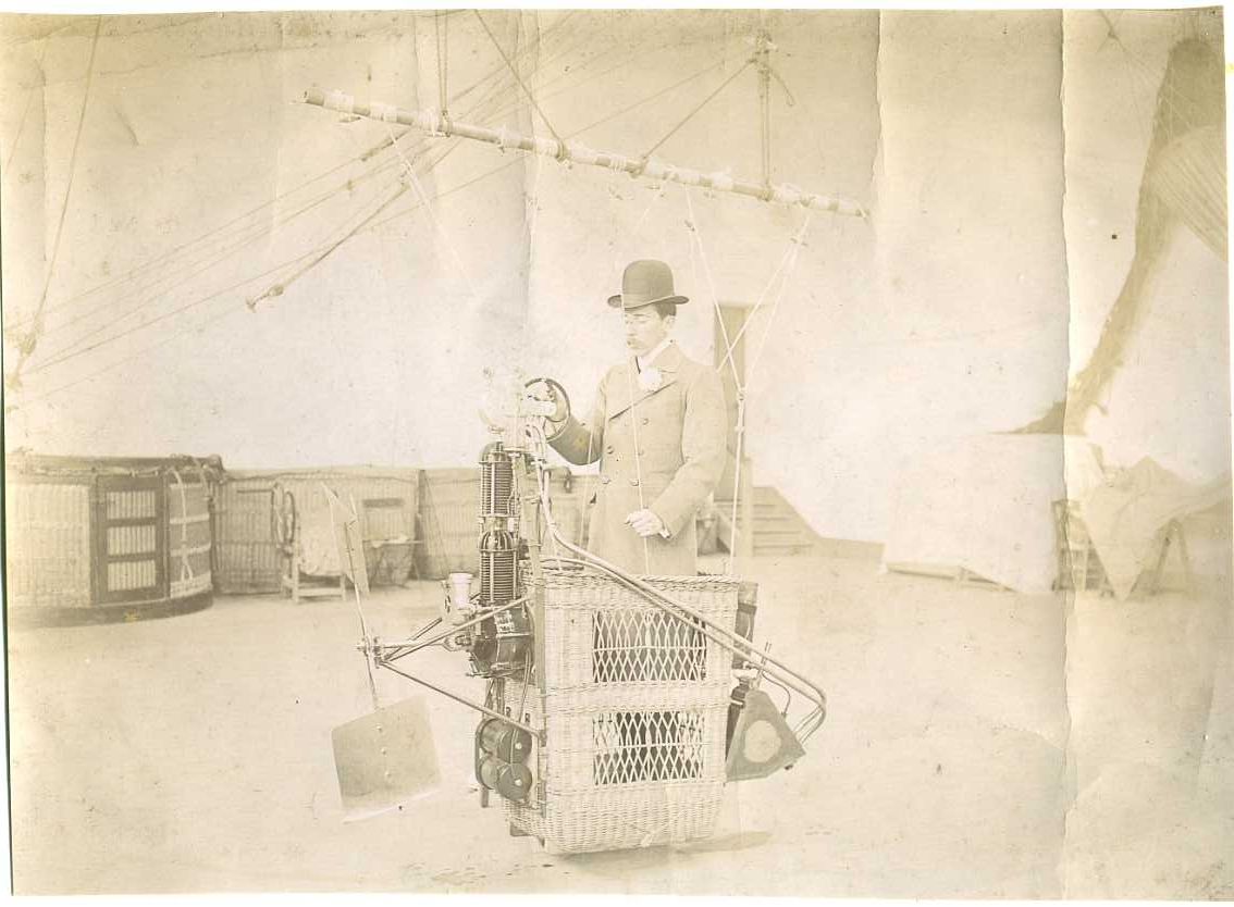 En man med kubb och rock står i en liten ballongkorg försedd med motor och propeller.