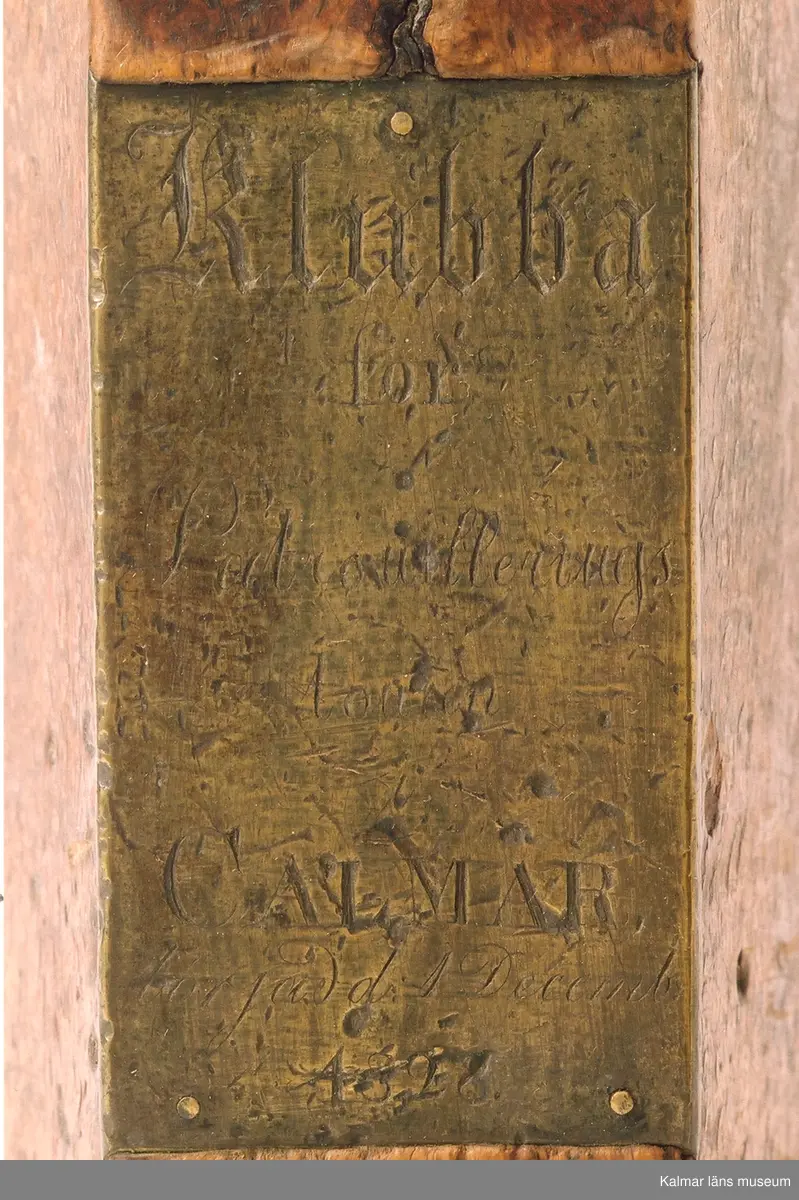 KLM 21316. Klubba, patrouilleringsklubba. Tillverkad av trä, masurbjörk och mässingsplåt. Femkantig klubba med på ena sidan graverad mässingsplåt med text: Klubba för Patrouilleringstouren i Calmar börjad d 1 Decemb 1828. Skaftet sönder, äldre lagning med järnnit.