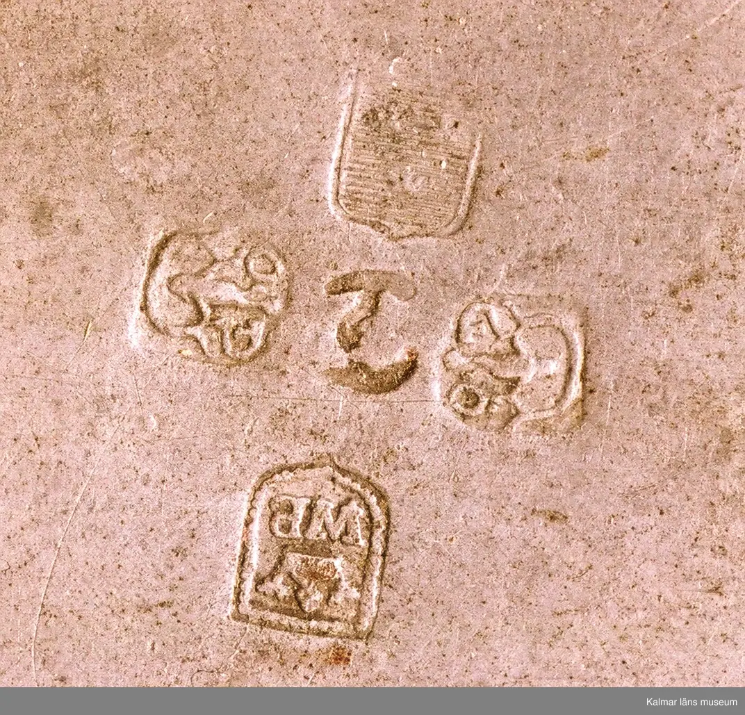 KLM 23468. Kallskål. Rund skål av tenn med två handtag. Runt omkring prydd med fågel- och blommotiv. Stämplar inuti botten: tre kronor (kontrollstämpel), W.B, med en krona över, Vimmerby (stadsstämpel), två stämplar G.Ö. för Gudmund Östling 1762 - 1790 (mästarstämpel), samt ett T, för 1777 (årsstämpel). Inristat, ägarmärkning?: EKS.