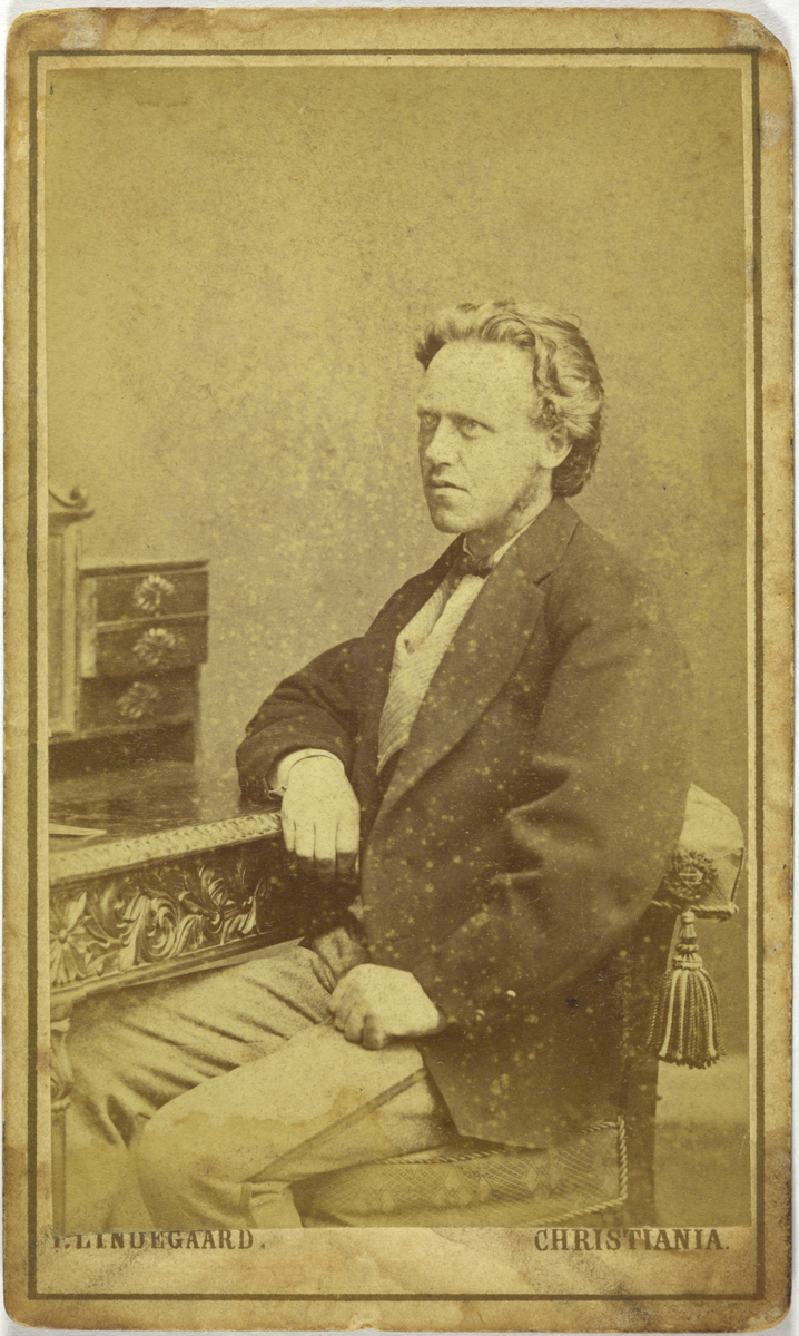 Atelierfoto av mann ved eit skrivebord. På baksida står det "Norheim?".