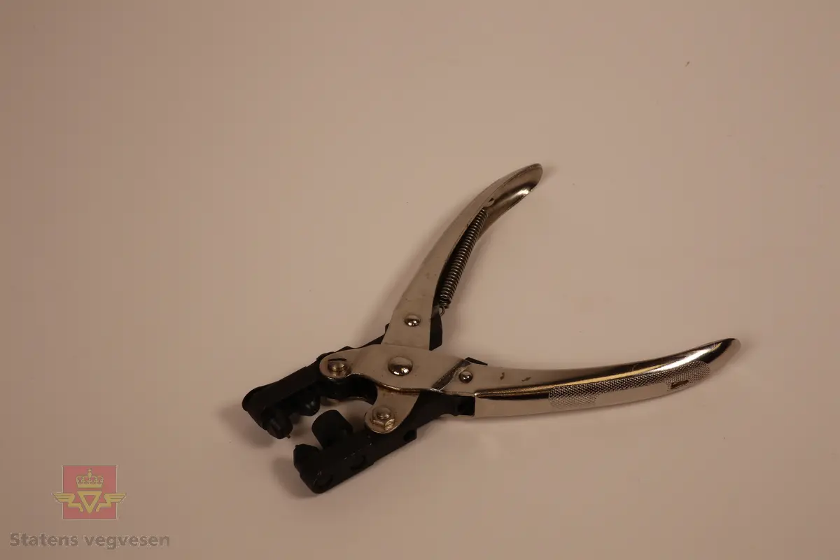 Verktøy til å reparere målebånd i metall i forskjellige bredder. inneholder skjøter i tre forskjellige bredder. 1cm, 1,3cm og 1,6cm. repareres ved å plassere en skjøt bak den knekte delen av et målebånd for å så nagle gjennom på hver side. Boksen er brun og åpnes ved å løfte av lokket. I  boksen finner man en tang for å feste nagler, metalskiver i tre størrelser til å repparere målebånd hvor noen er pakket inn i fettpapir. Det er også en liten grønn boks med en brun papirpose som inneholder nagler til tangen. 