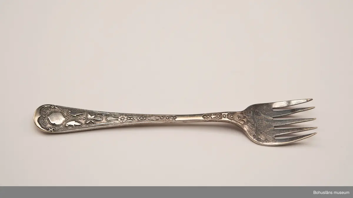 Sardingaffel av nysilver. U-format blad med fem klor, smalt skaft som vidgas upptill. Ornament i form av graverad sardin på bladet och ciselerade växtornament på skaftet. Baksida med graverade initialerna M L (Maria Lundbäck) samt otydliga stämplar.
Sliten.