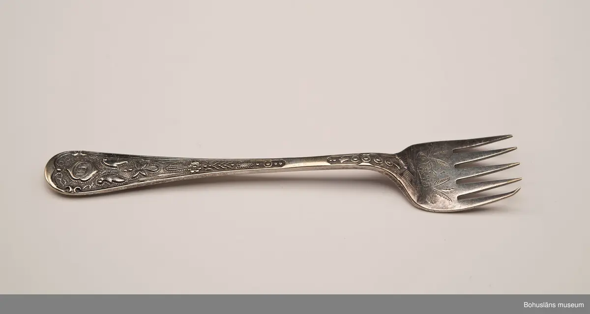 Sardingaffel av nysilver. U-format blad med fem klor, smalt skaft som vidgas upptill. Ornament i form av graverad sardin på bladet och ciselerade växtornament på skaftet. Skaftet med graverade initialerna M L (Maria Lundbäck). Slät baksida med otydliga stämplar.
Sliten.
