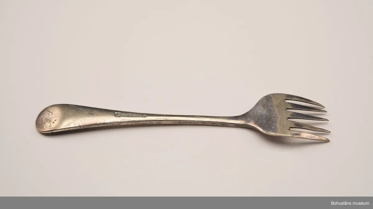 Sardingaffel av nysilver. U-format blad med fem klor, smalt skaft som vidgas upptill. Ornament i form av graverad sardin på bladet och ciselerade växtornament på skaftet. Baksida med graverade initialerna M L (Maria Lundbäck) samt otydliga stämplar.
Sliten.