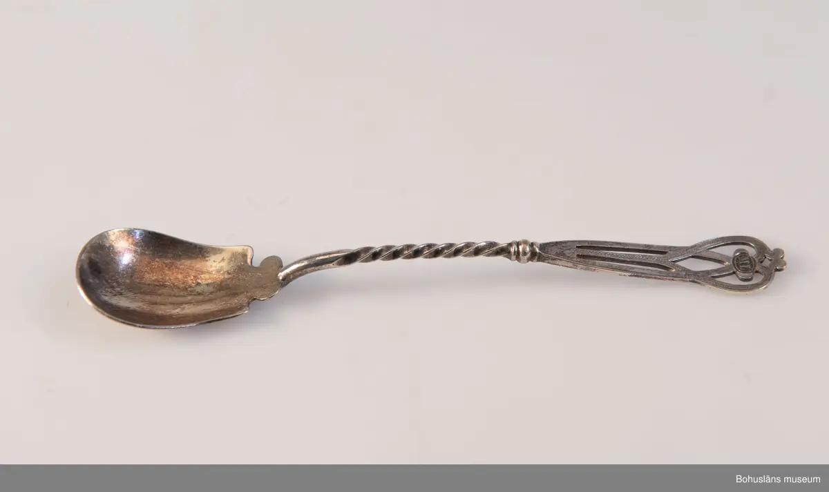 Serveringsked av nysilver. Ovalt blad, vridet skaft med genombrutet avslut i jugendslingor med punsad dekor och graverade initialerna M L (Maria Lundbäck). Slät baksida med otydliga stämplar.