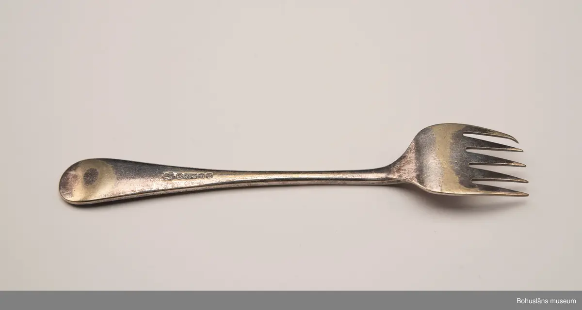 Sardingaffel av nysilver. U-format blad med fem klor, smalt skaft som vidgas upptill. Ornament i form av graverad sardin på bladet och ciselerade växtornament på skaftet. Skaftet med graverade initialerna M L (Maria Lundbäck). Slät baksida med otydliga stämplar.
Sliten.