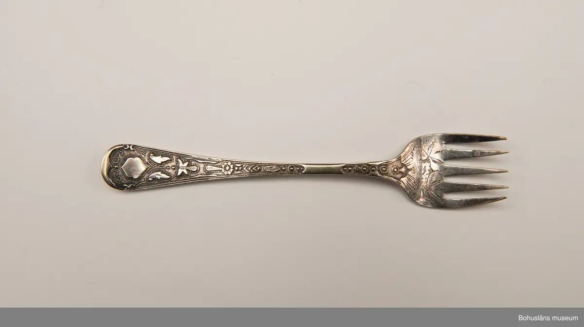 Sardingaffel av nysilver. U-format blad med fem klor, smalt skaft som vidgas upptill. Ornament i form av graverad sardin på bladet och ciselerade växtornament på skaftet. Baksida med graverade initialerna M L (Maria Lundbäck) samt otydliga stämplar.
Sliten.