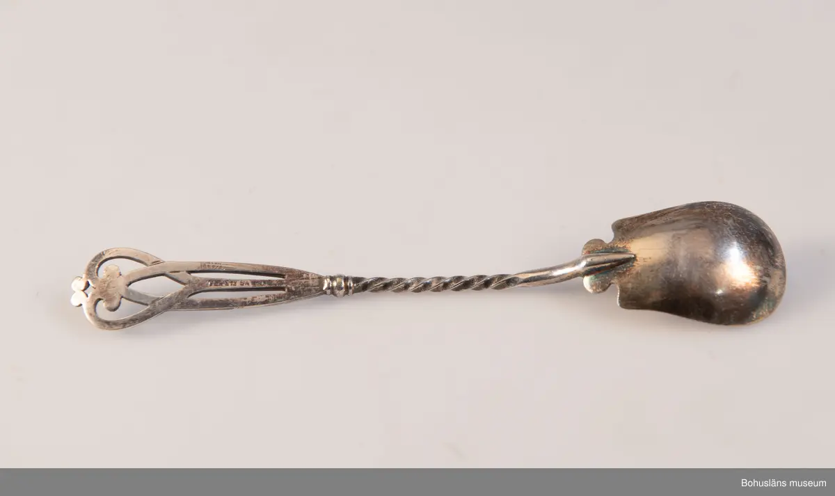 Serveringsked av nysilver. Ovalt blad, vridet skaft med genombrutet avslut i jugendslingor med punsad dekor och graverade initialerna M L (Maria Lundbäck). Slät baksida med otydliga stämplar.