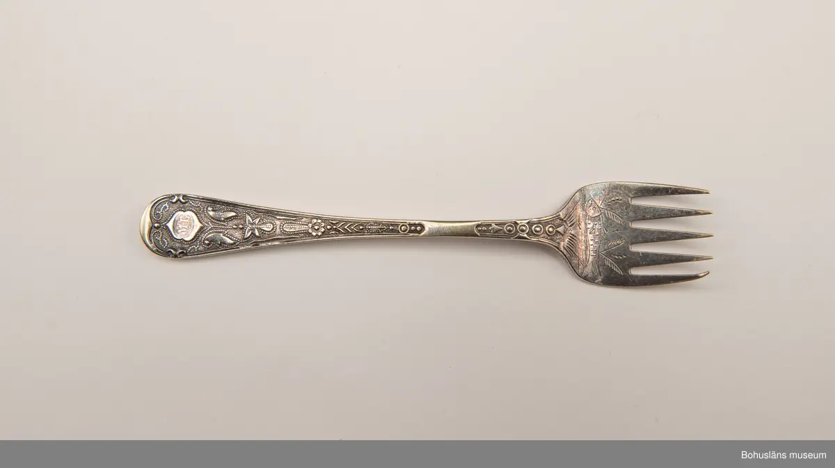 Sardingaffel av nysilver. U-format blad med fem klor, smalt skaft som vidgas upptill. Ornament i form av graverad sardin på bladet och ciselerade växtornament på skaftet. Skaftet med graverade initialerna M L (Maria Lundbäck). Slät baksida med otydliga stämplar.
Sliten.