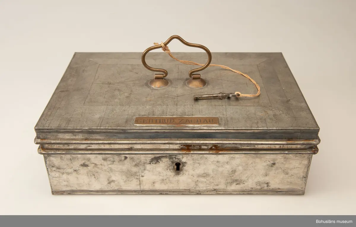 Rektangulär plåtlåda med lock. Handtag och nyckelhål. På lockets utsida en mässngsplåt med ingraverade texten GERTRUD ZACHAU.
Tillhörande nyckel.
Vid förvärvet innehöll skrinet fotogafier, se UMFA55947 och handlingar, se BMEA 488.