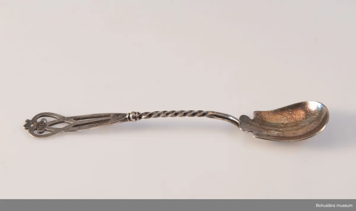 Serveringsked av nysilver. Ovalt blad, vridet skaft med genombrutet avslut i jugendslingor med punsad dekor och graverade initialerna M L (Maria Lundbäck). Slät baksida med otydliga stämplar.