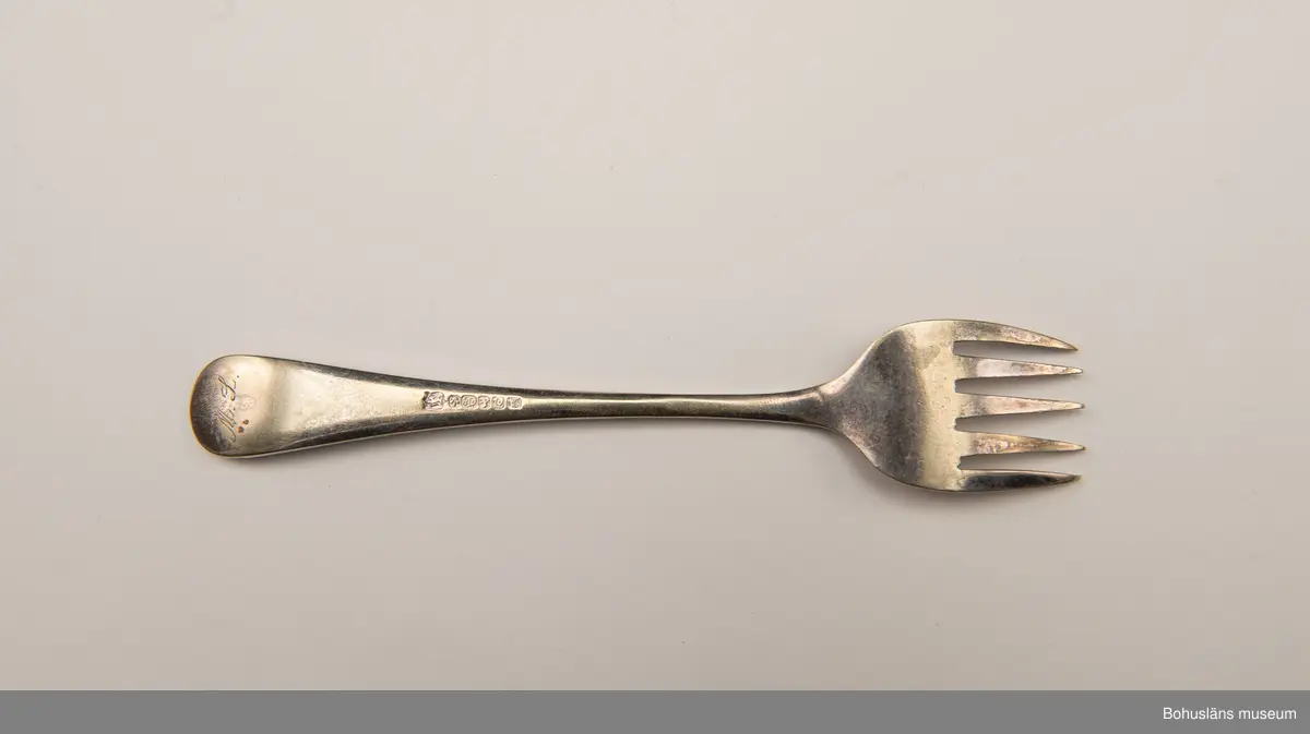 Sardingaffel av nysilver. U-format blad med fem klor, smalt skaft som vidgas upptill. Ornament i form av graverad sardin på bladet och ciselerade växtornament på skaftet. Baksida med graverade initialerna M L (Maria Lundbäck) samt otydliga stämplar.
Sliten.