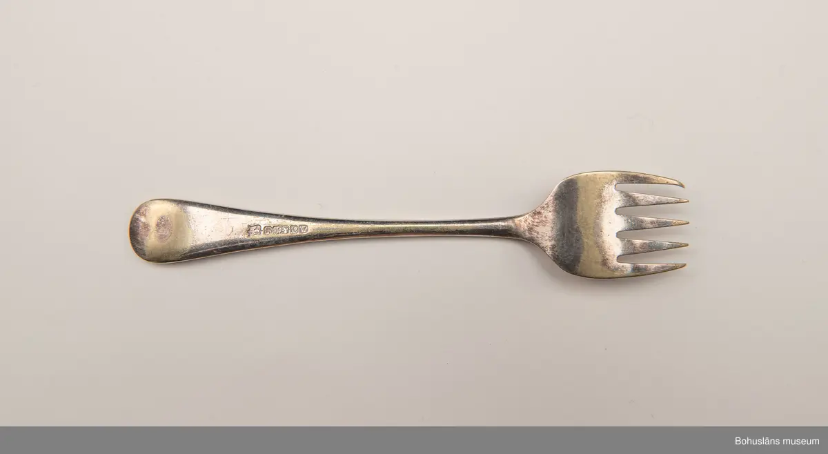 Sardingaffel av nysilver. U-format blad med fem klor, smalt skaft som vidgas upptill. Ornament i form av graverad sardin på bladet och ciselerade växtornament på skaftet. Skaftet med graverade initialerna M L (Maria Lundbäck). Slät baksida med otydliga stämplar.
Sliten.