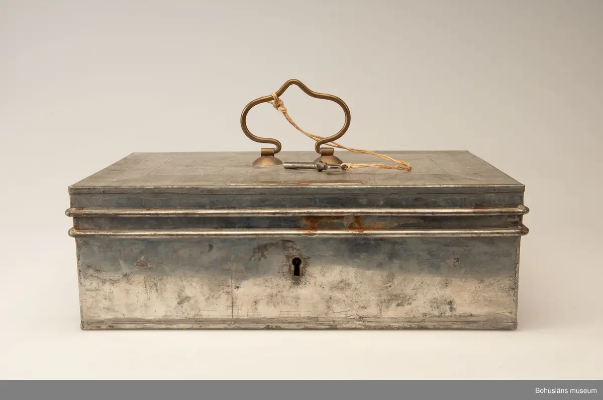 Rektangulär plåtlåda med lock. Handtag och nyckelhål. På lockets utsida en mässngsplåt med ingraverade texten GERTRUD ZACHAU.
Tillhörande nyckel.
Vid förvärvet innehöll skrinet fotogafier, se UMFA55947 och handlingar, se BMEA 488.